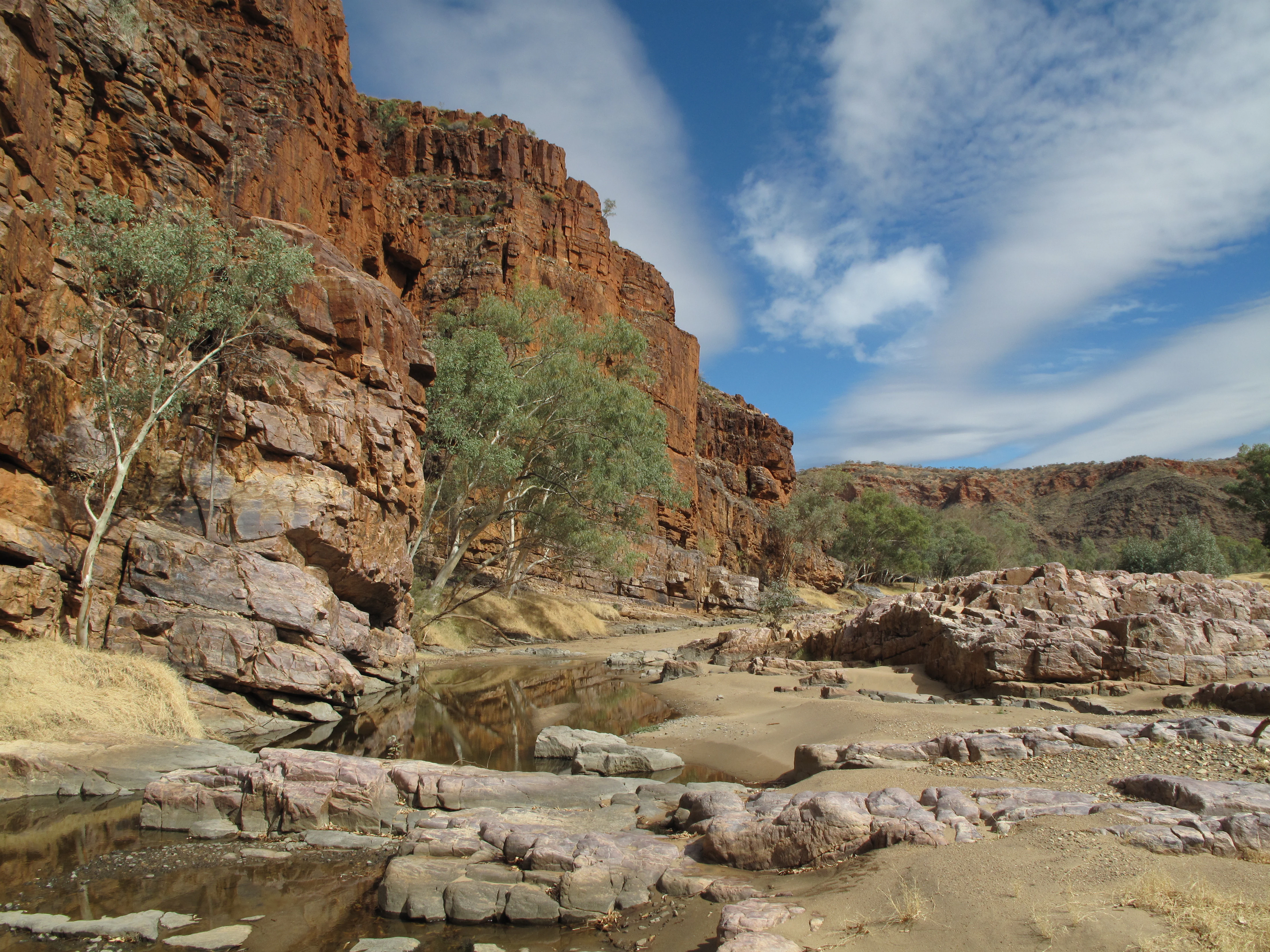 A landscape view of reddish-brown rocky cliffs and a dry riverbed with shallow pools of water and sparse green trees under a blue sky with white clouds.