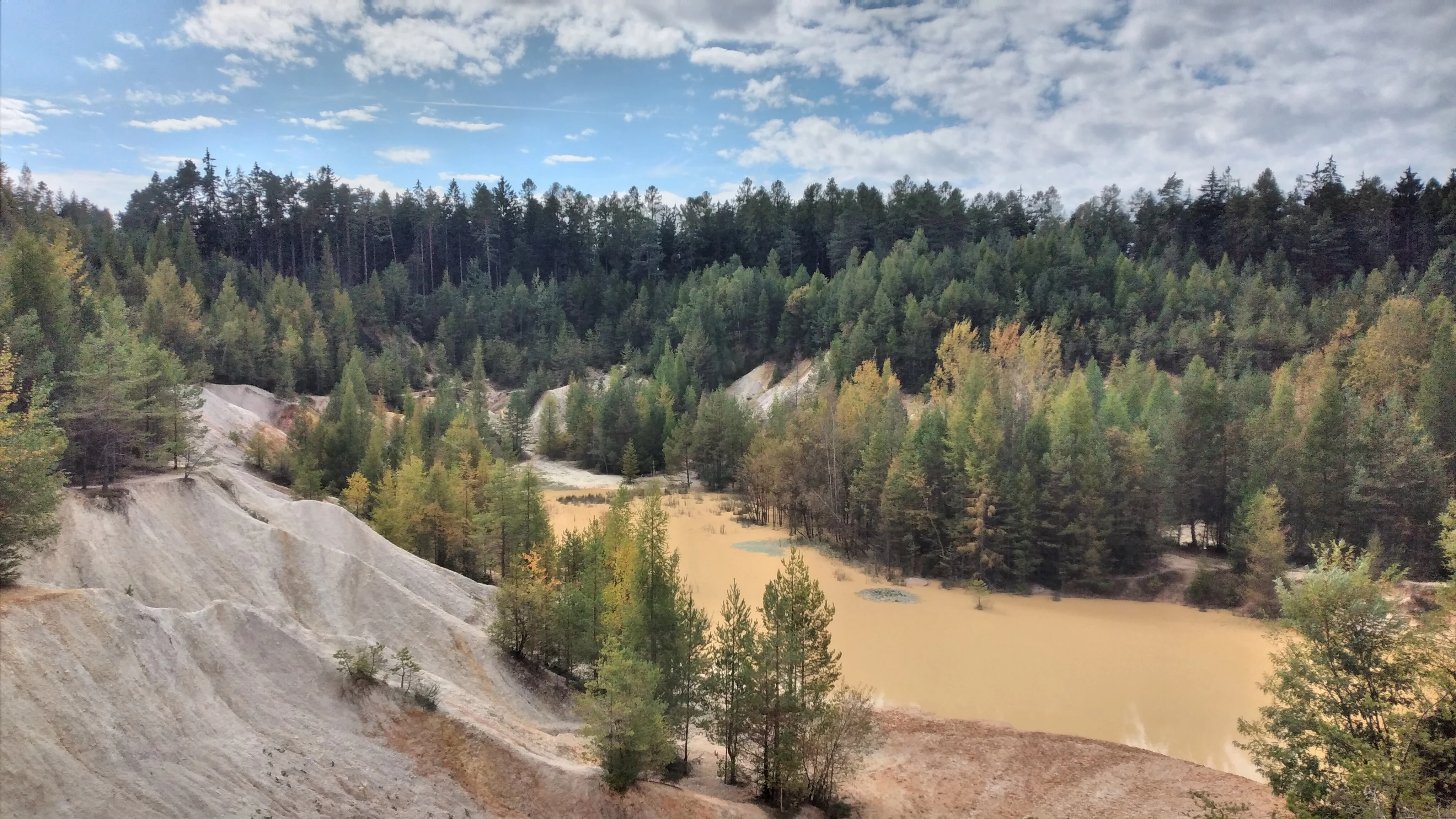 A high-angle view of a former quarry featuring a light-colored sandy basin and a shallow, murky pond surrounded by dense coniferous and deciduous forest under a partly cloudy sky.
