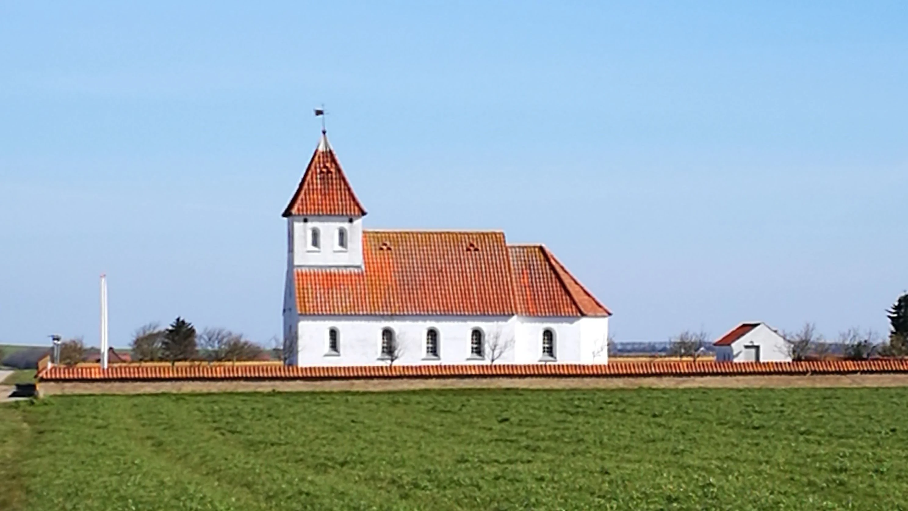 A white church with a red tiled roof and a small tower, surrounded by a low brick wall, standing in a green field under a clear blue sky.