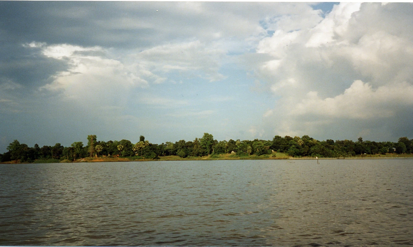 A wide view of a calm lake with a dense tree-lined shoreline in the distance under a partly cloudy sky.