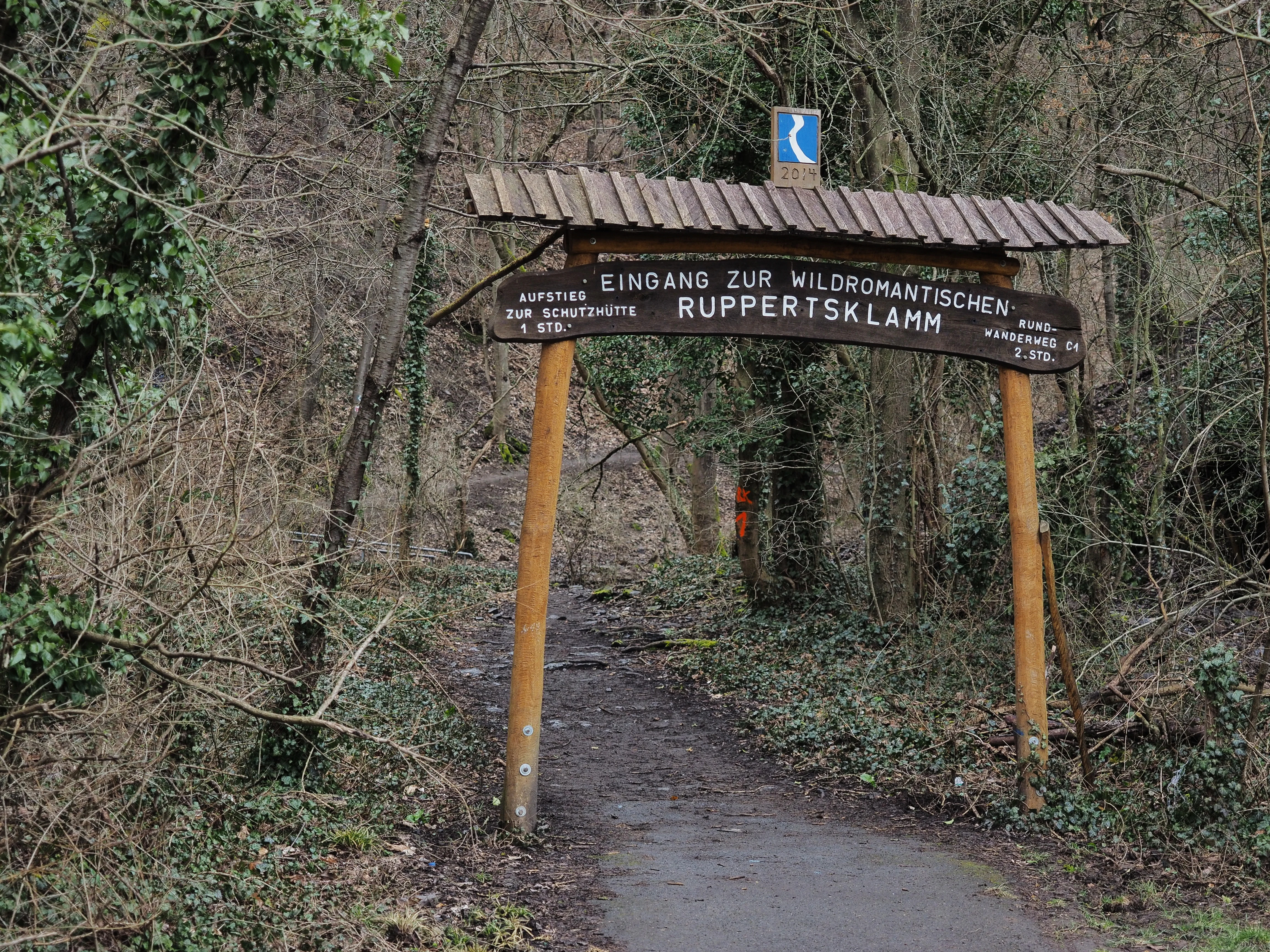 A rustic wooden arched entrance sign marked 'Eingang zur wildromantischen Ruppertsklamm' stands at the beginning of a forest trail.