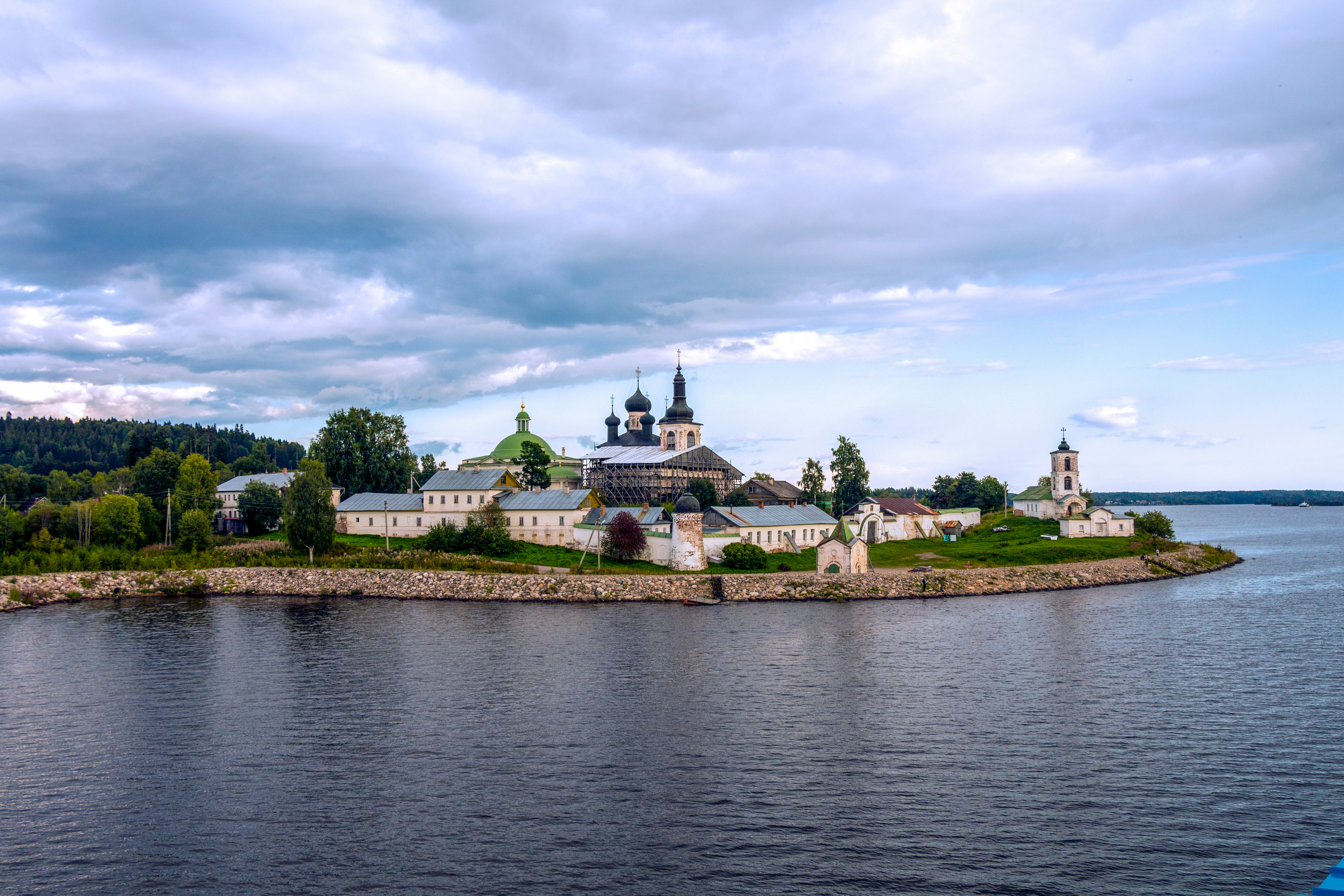 A distant view of the Goritsky Monastery complex with white buildings and dark domes, located on a grassy shore next to a wide river under a cloudy sky.