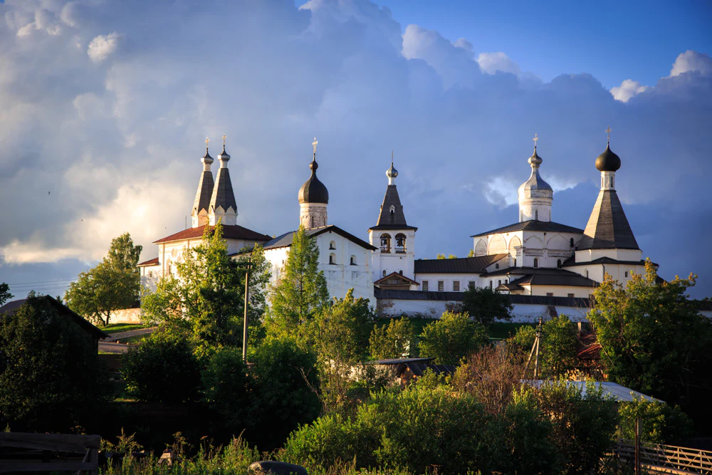 A group of white church buildings with onion domes sits on a gentle hill at sunset, surrounded by green trees and foliage under a sky filled with large, puffy clouds.