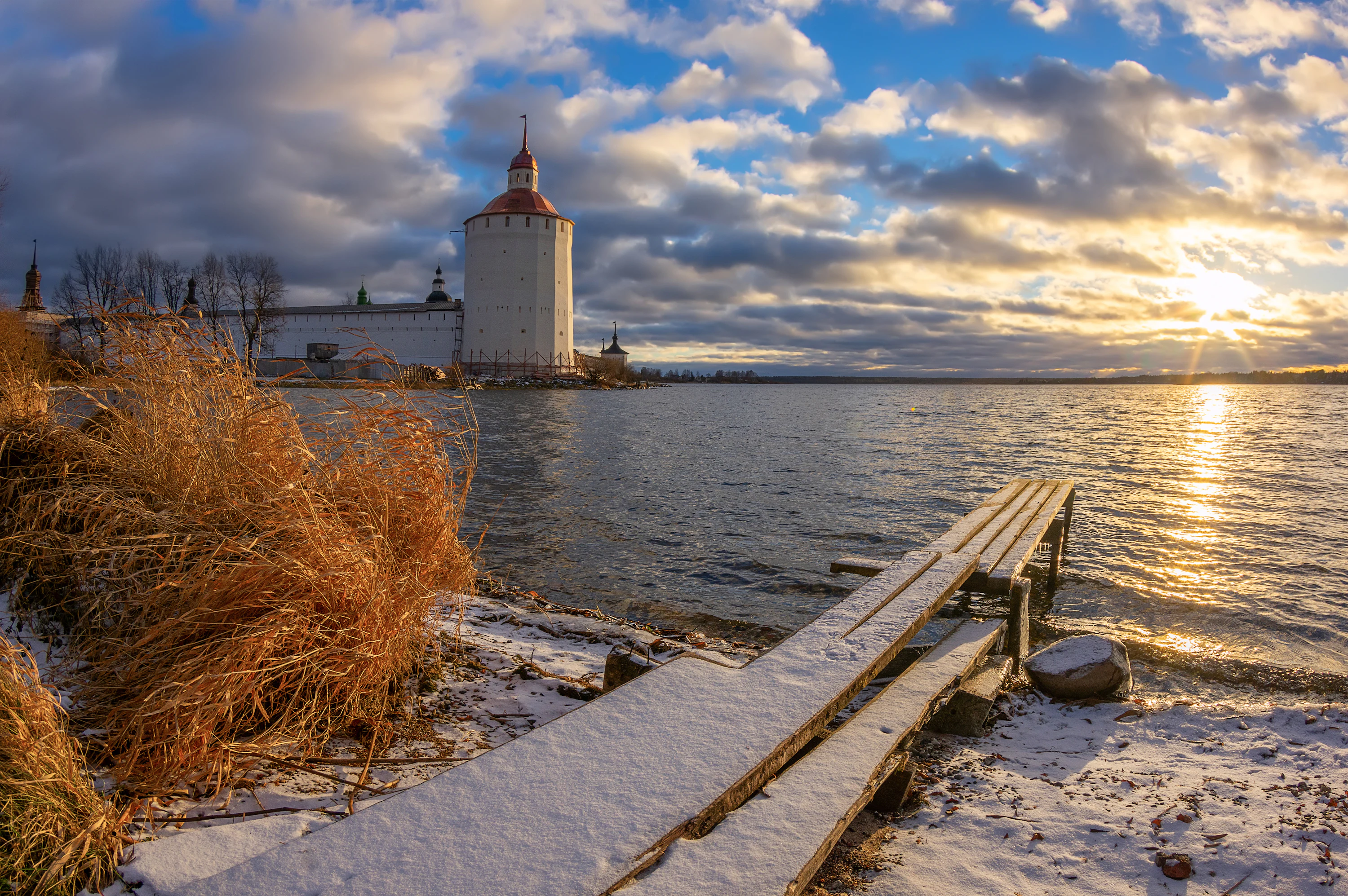 A snowy, wooden pier leads into the water of a lake at sunset, with the white walls and towers of a historic stone monastery visible on the far shore under a dramatic, cloudy sky.