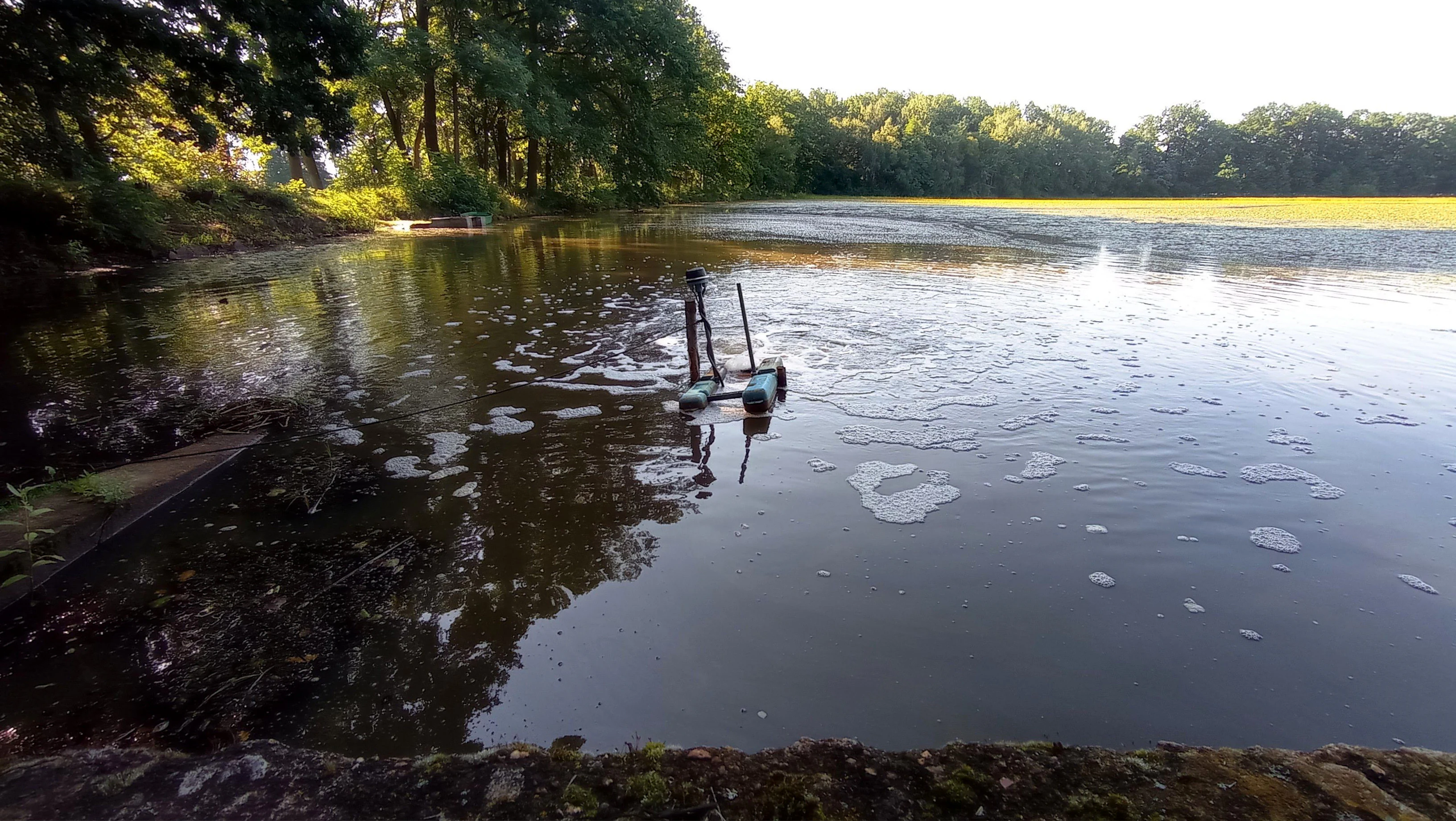 A calm, dark-water pond surrounded by trees, with a small mechanical aerator creating ripples in the center.