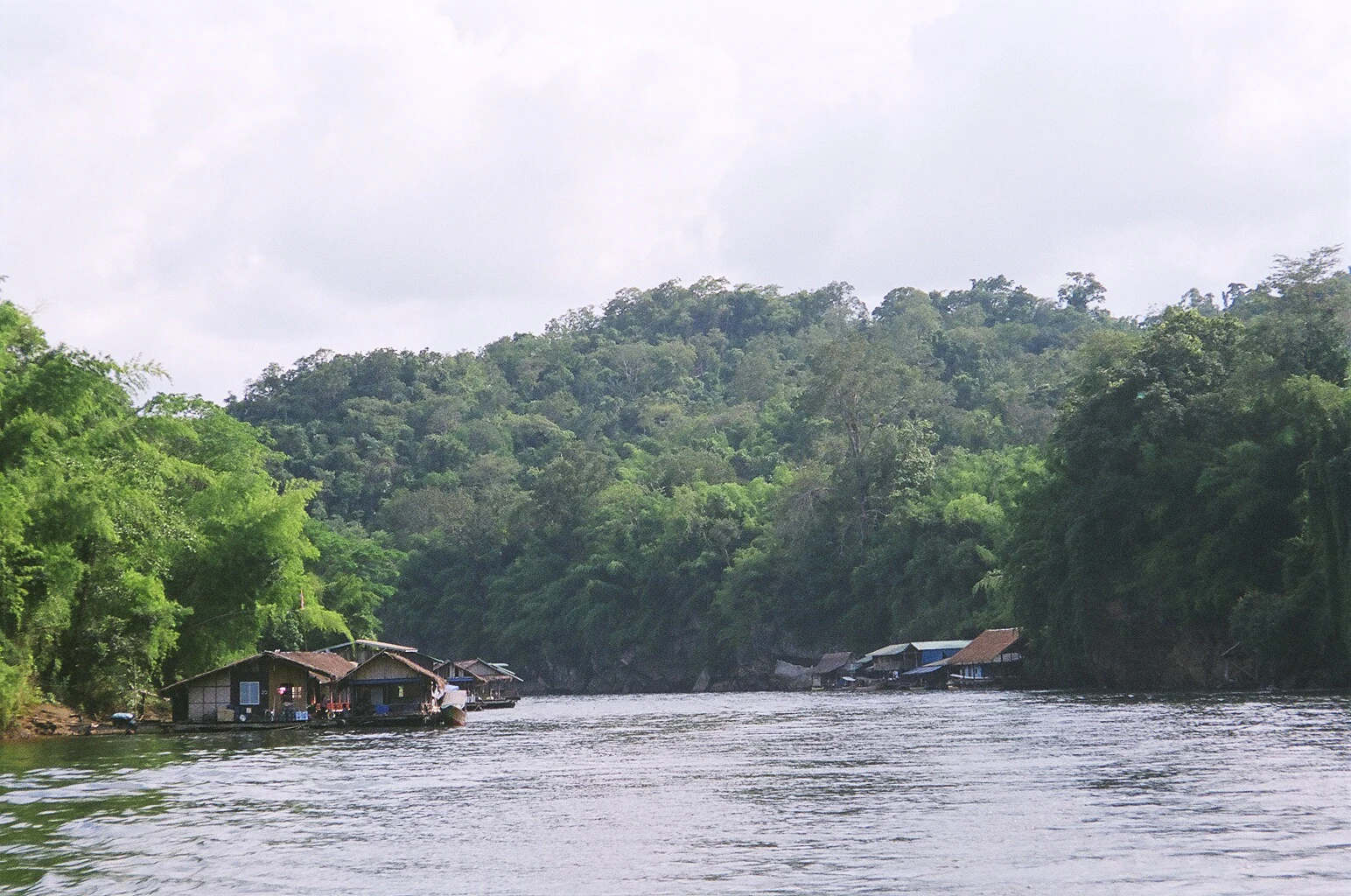 A wide river with calm water bordered by dense green forests and small riverfront structures under a partly cloudy sky.