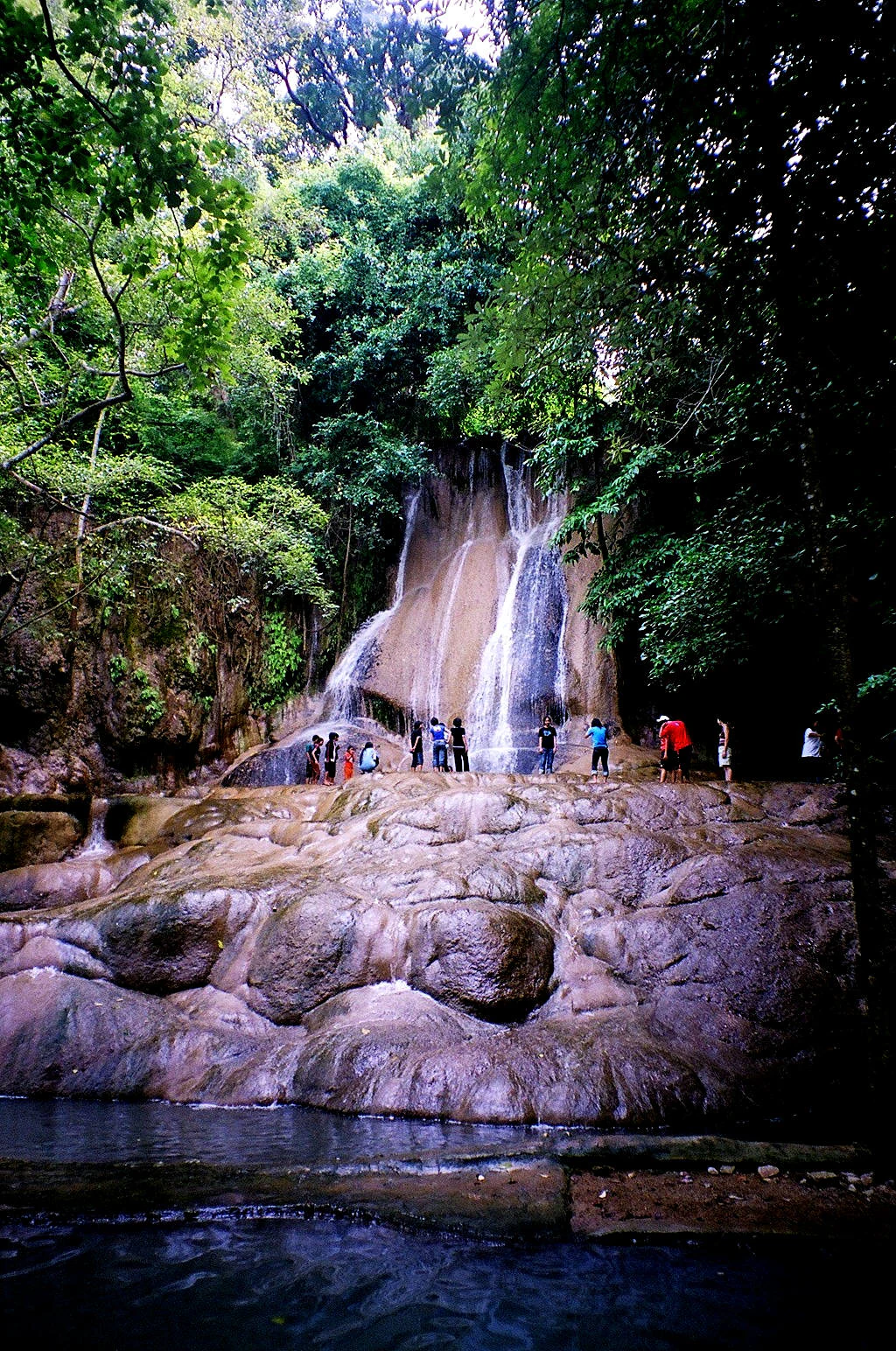 Waterfall cascading over rocky terrain with lush green forest surrounding it and people standing at the base near a pool of water