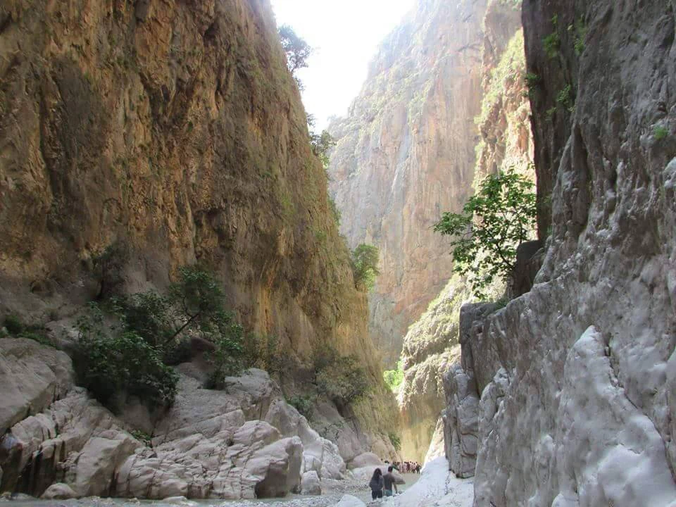 Steep rocky canyon walls with a narrow stream at the bottom and small group of people walking in the distance