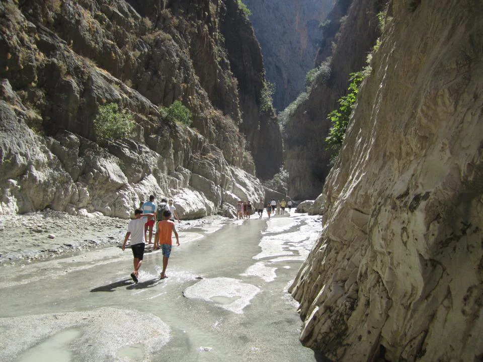 Two people walking on a dry riverbed in a deep canyon with rocky cliffs on both sides