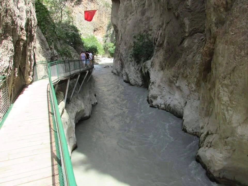 Rocky canyon with a flowing river, wooden boardwalk with green railing on left, and people walking along the path