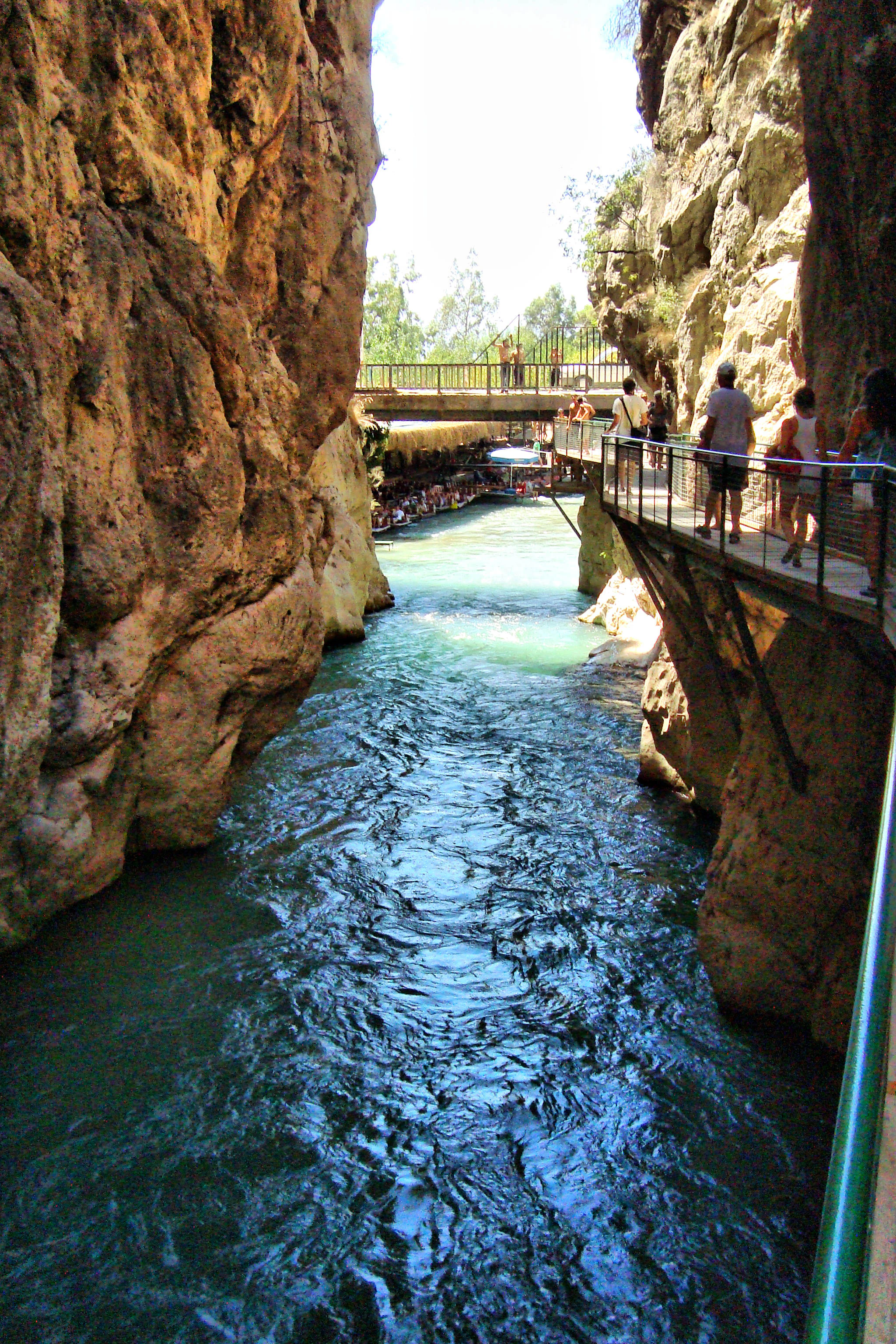 River flowing through a narrow canyon with steep rocky walls, metal walkway with visitors on the right, and a bridge in the background