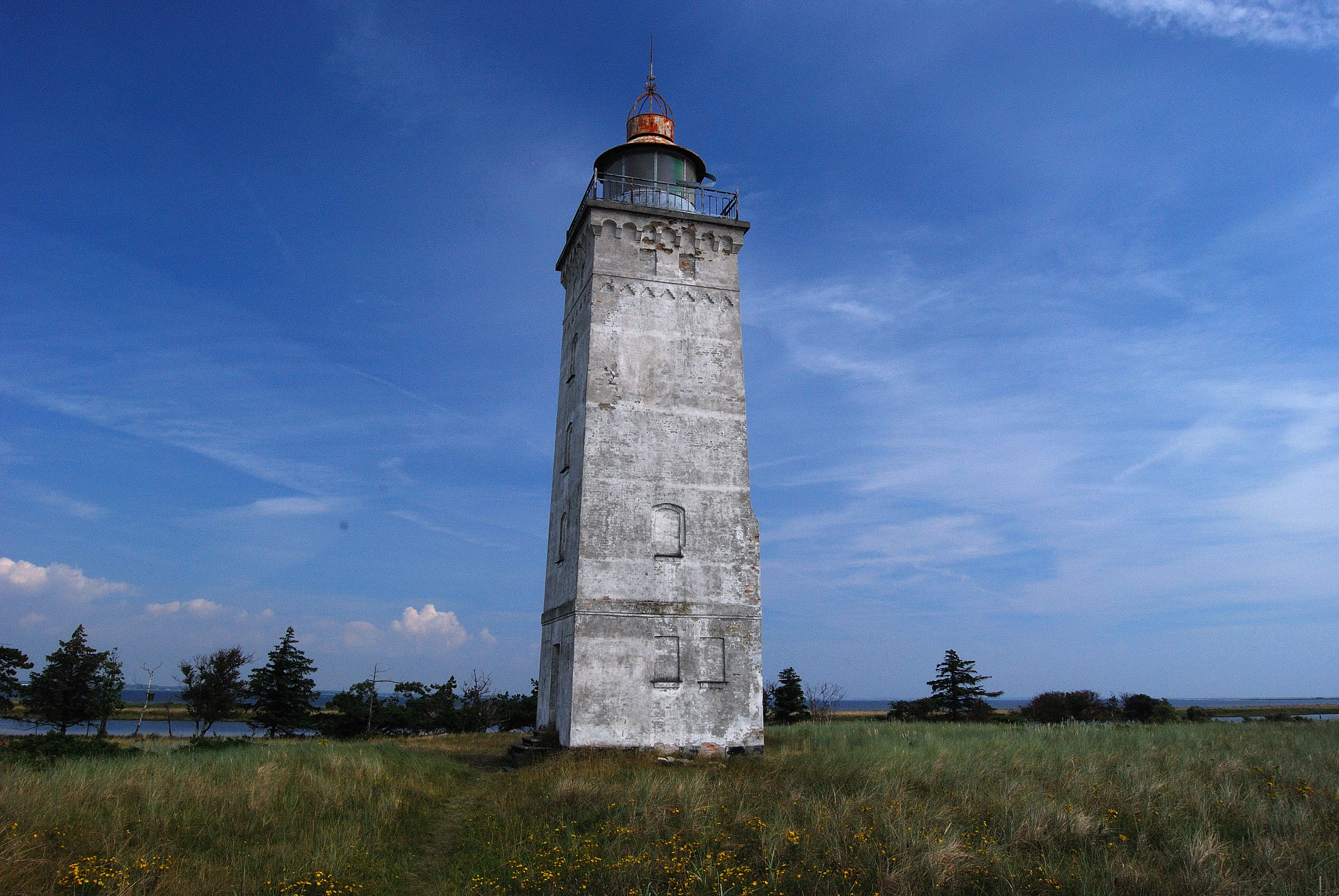 Un faro cuadrado de piedra blanca y desgastada se alza en un campo costero cubierto de hierba bajo un cielo azul despejado.