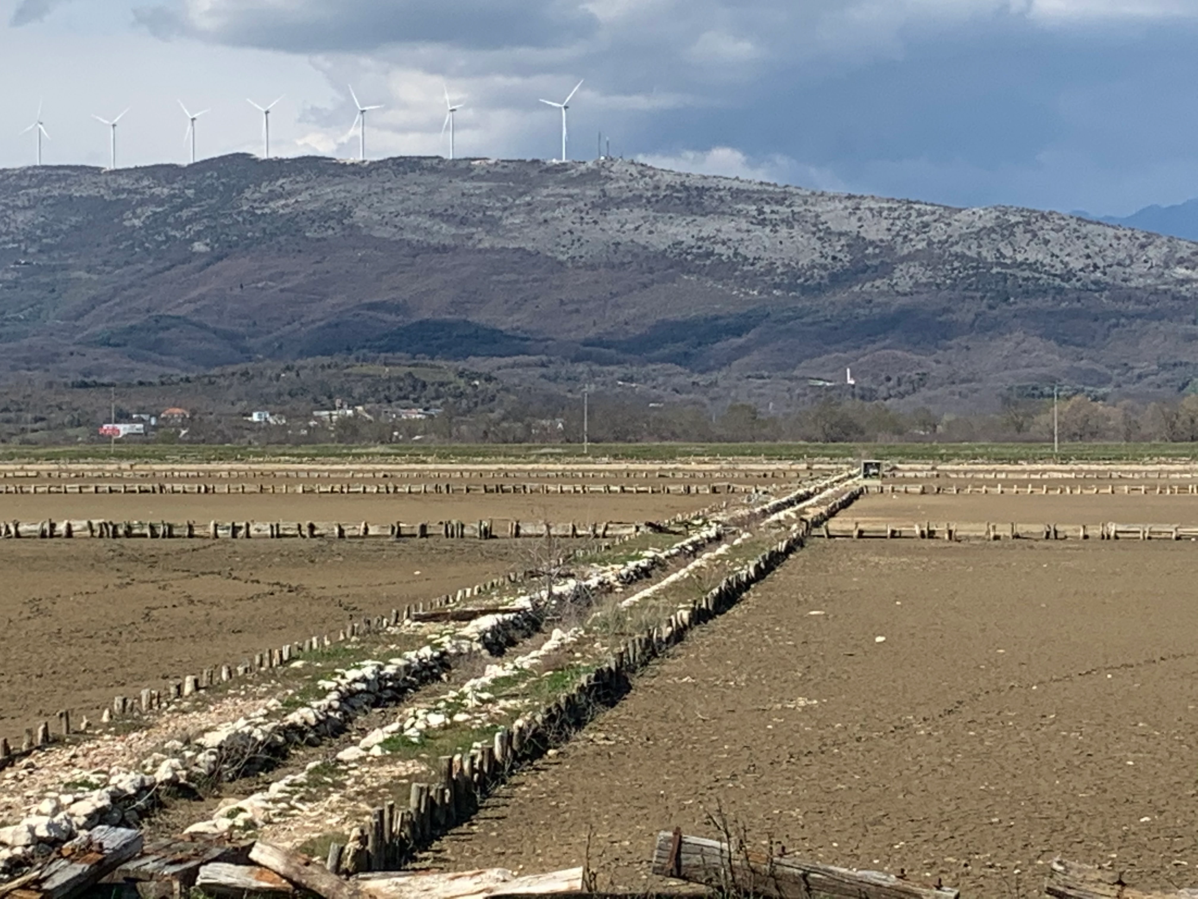 A wide view of the dry, brown salt pan fields at Saline Ulcinj, with wooden stake borders and low hills topped with wind turbines in the distance under a cloudy sky.