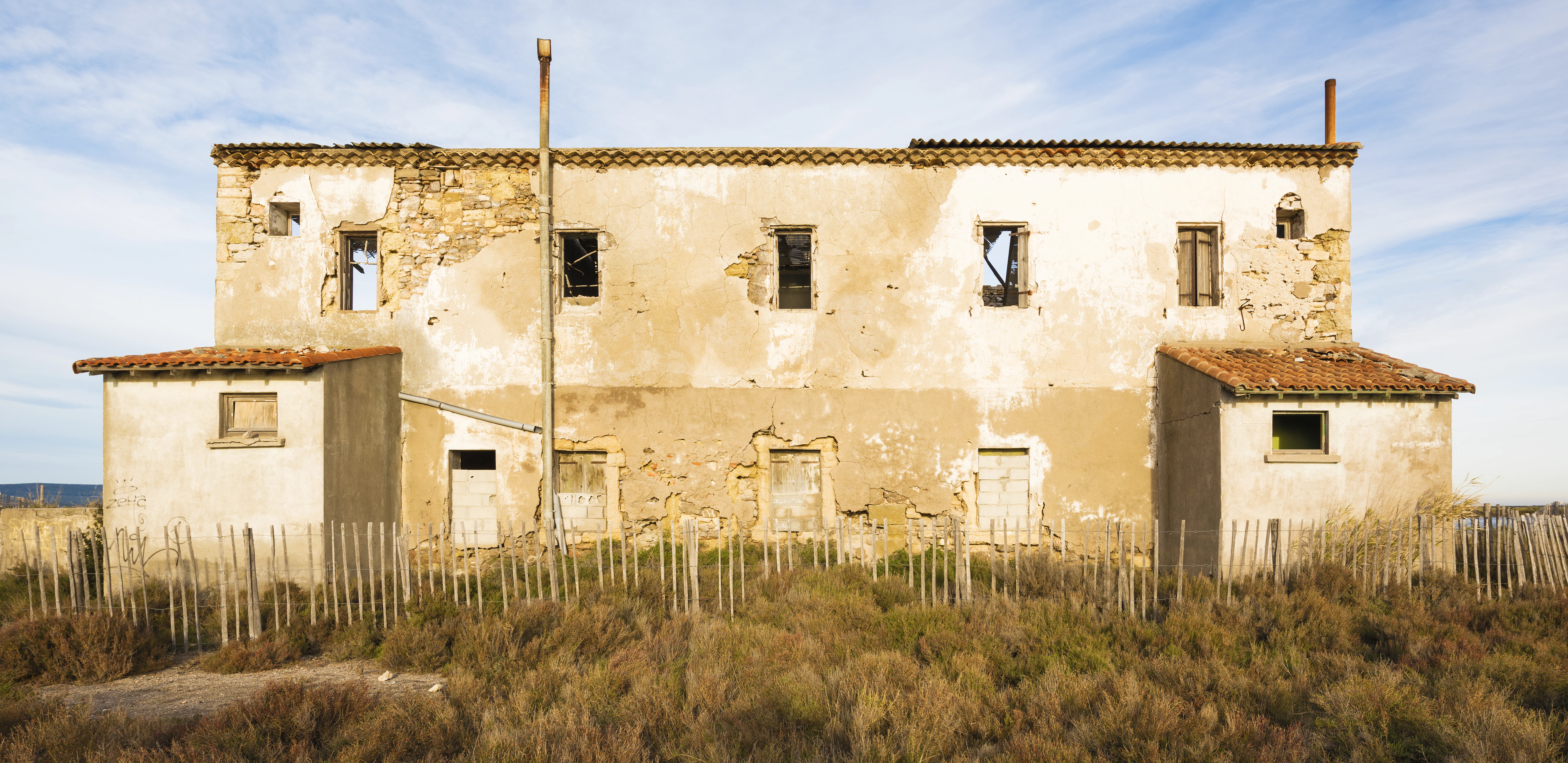 A weathered, abandoned two-story building with beige stucco walls, exposed stonework, and tiled roofs, situated behind a low wooden picket fence in a grassy area.