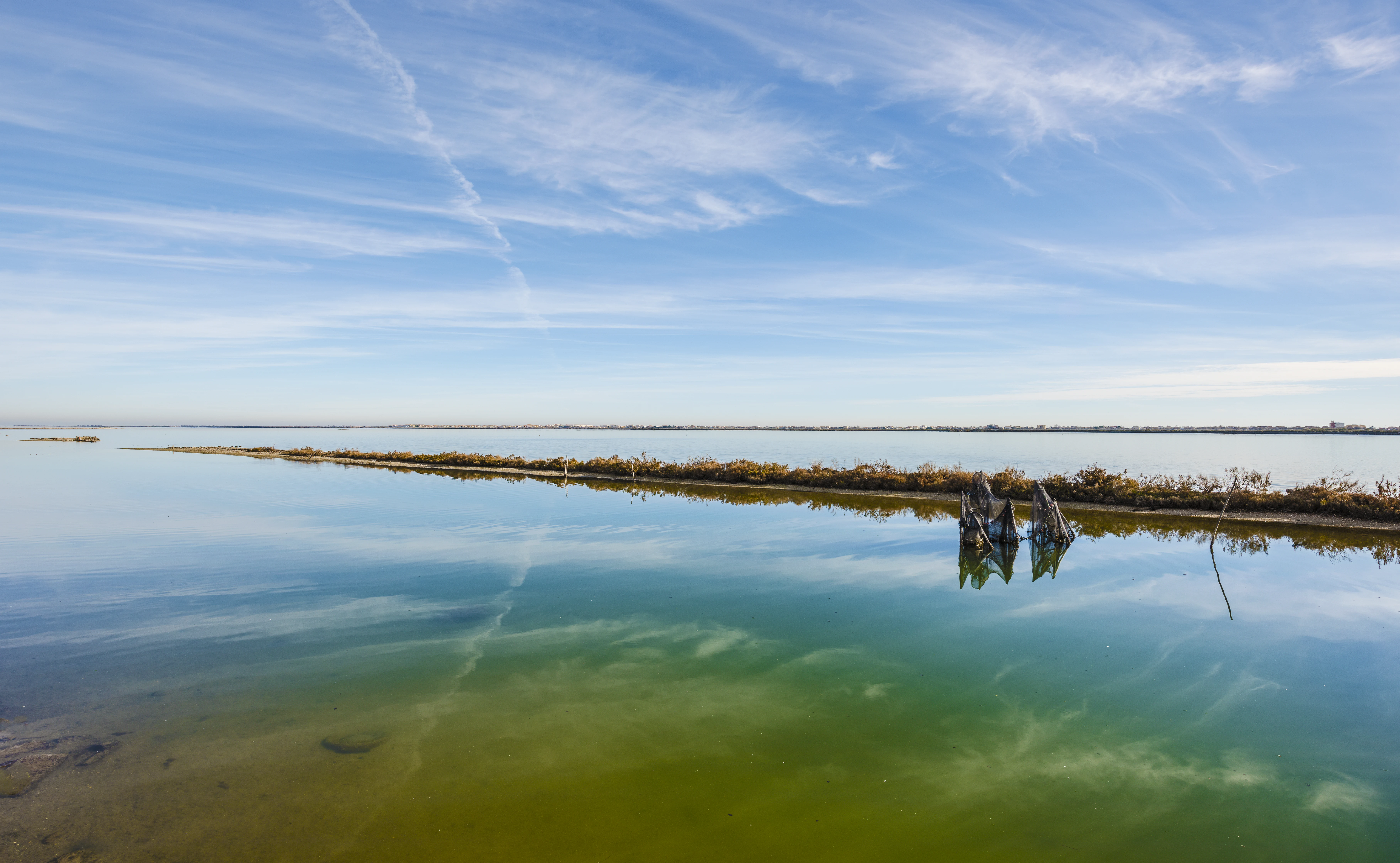 A wide, calm, shallow body of water reflecting a bright blue sky with wispy clouds, featuring a low, thin land strip covered in sparse vegetation and small wooden pilings in the water.