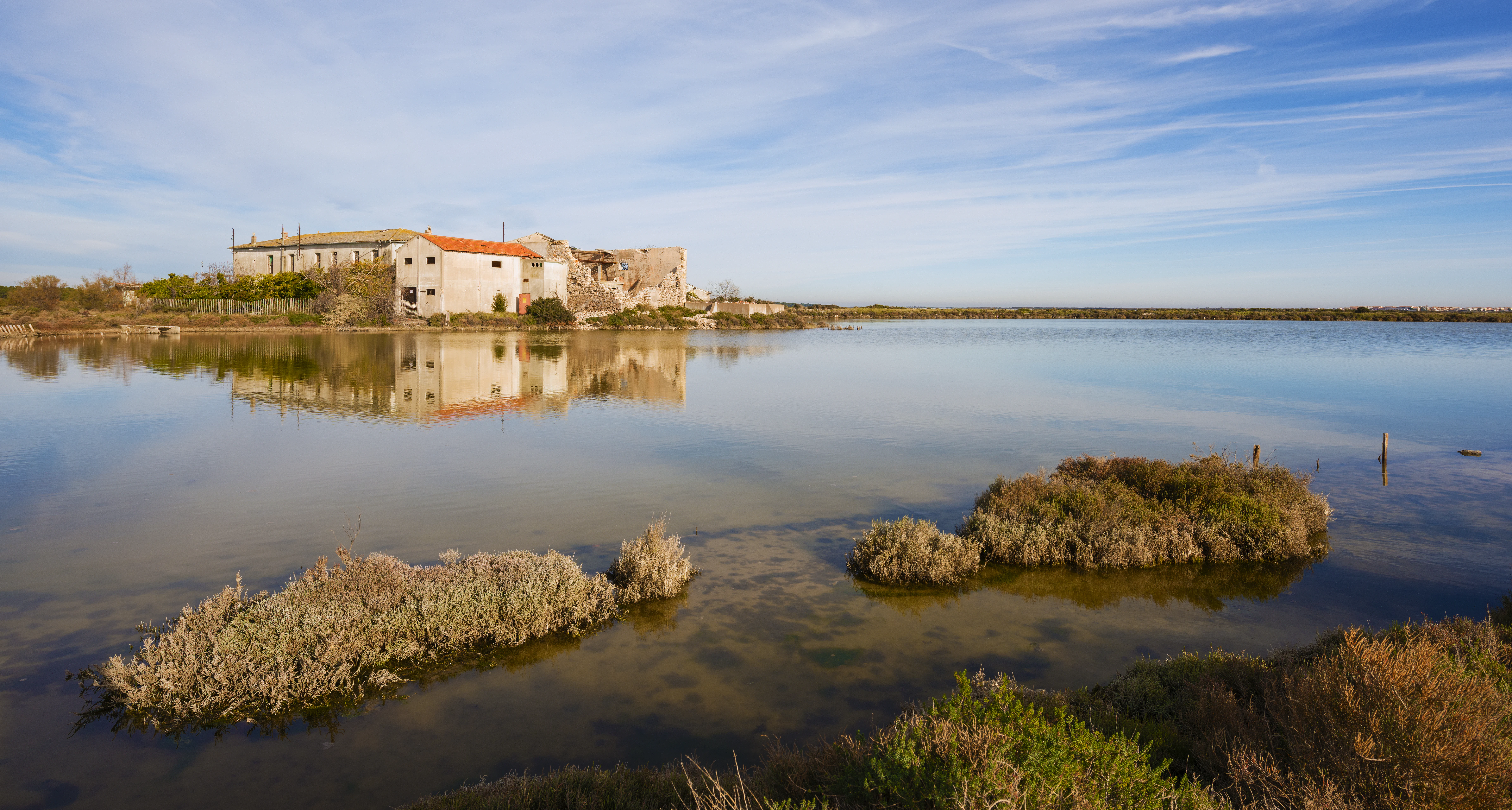 A wide, eye level shot of a cluster of weathered, low-rise stone buildings sitting on the shore of a shallow, reflective wetland area, with low-lying coastal vegetation in the foreground and a clear blue sky above.