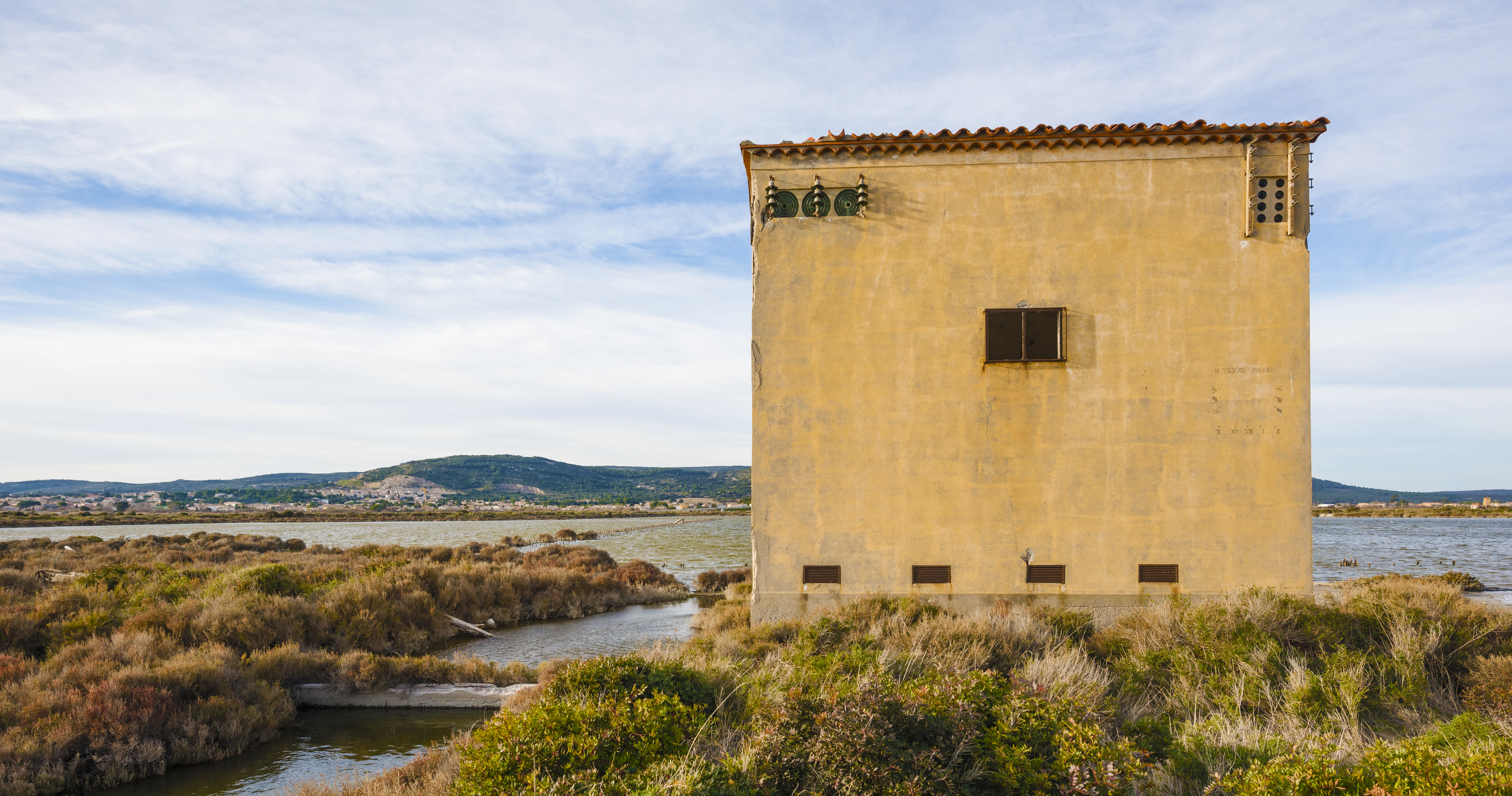 A square, tan-colored industrial building stands in the center of a salt marsh, surrounded by low-lying vegetation and water with distant hills on the horizon.