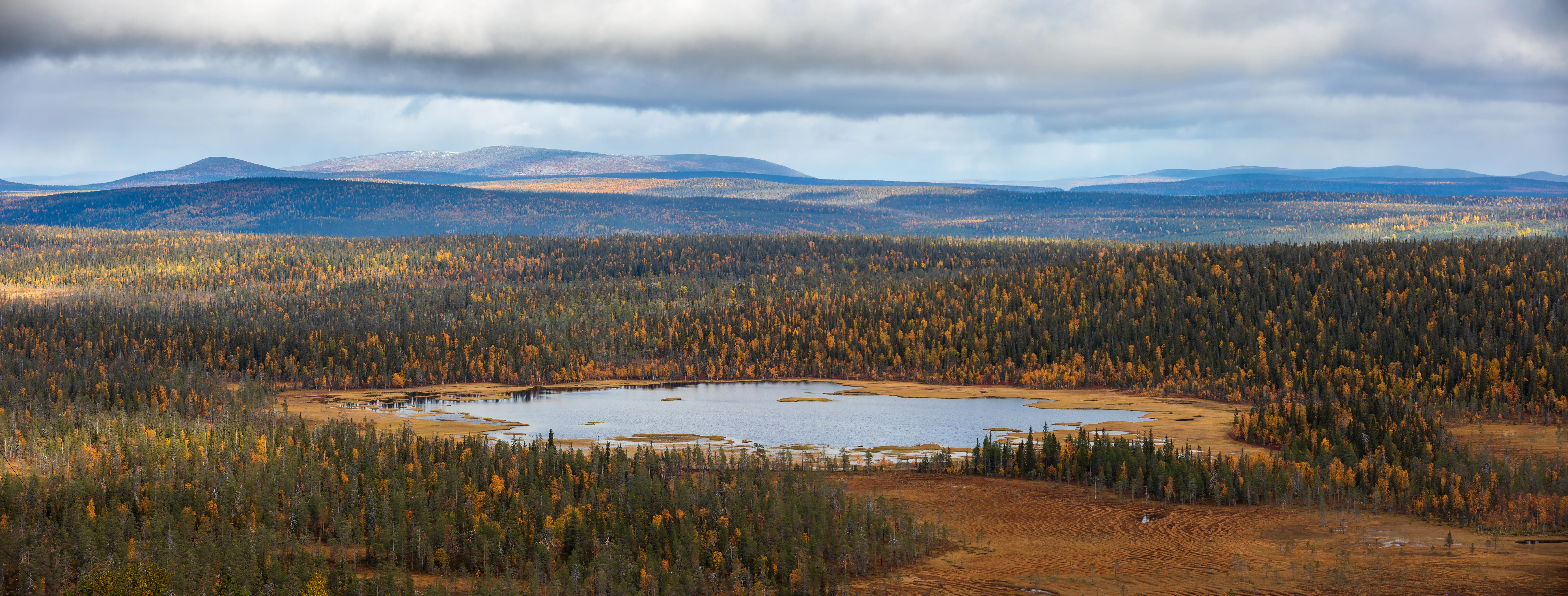 Wide aerial view of a lake surrounded by autumn-colored forest with rolling hills under a cloudy sky