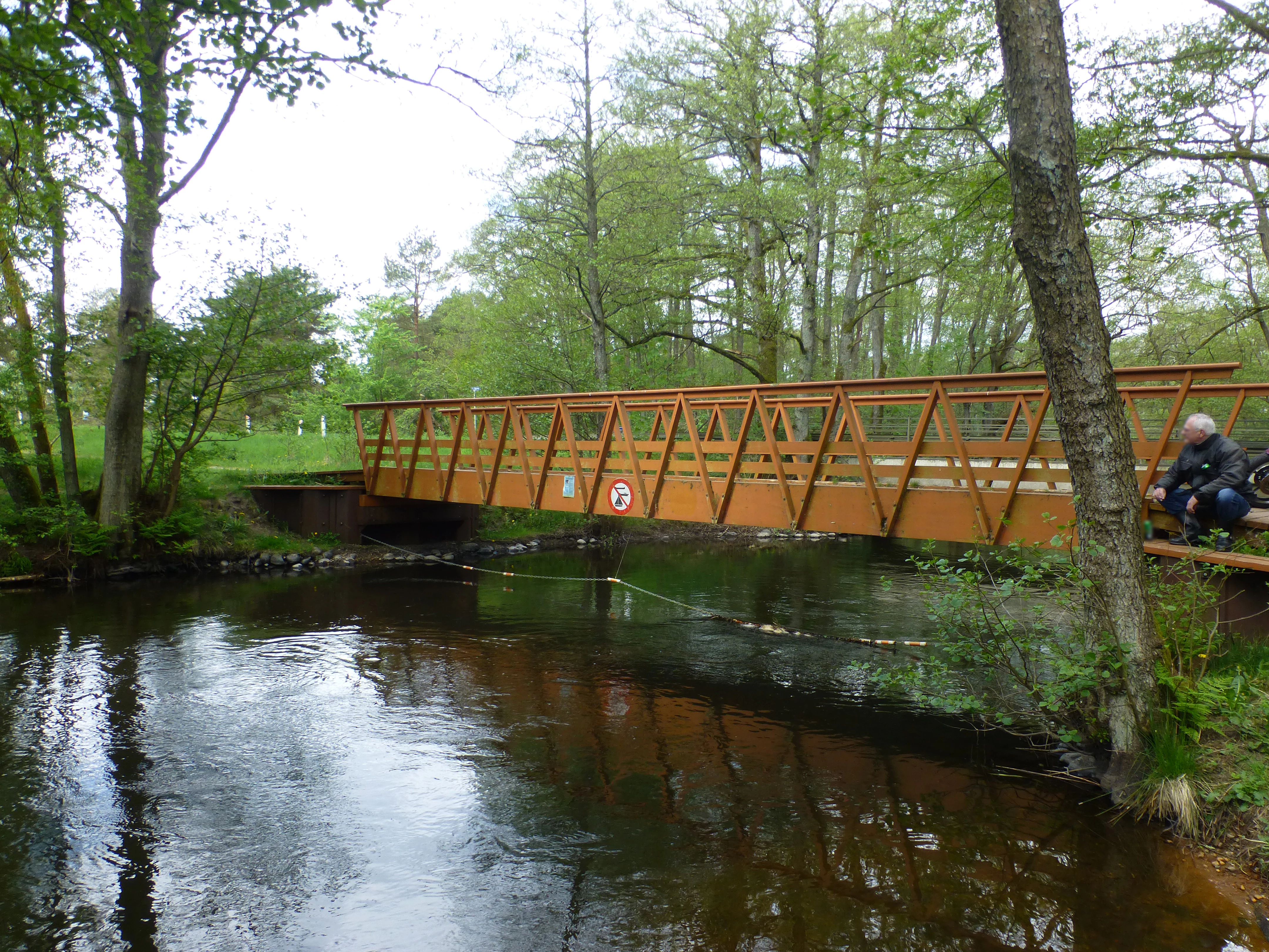A wooden truss bridge crosses over the calm, dark waters of the Salten Aa river, surrounded by lush deciduous trees.