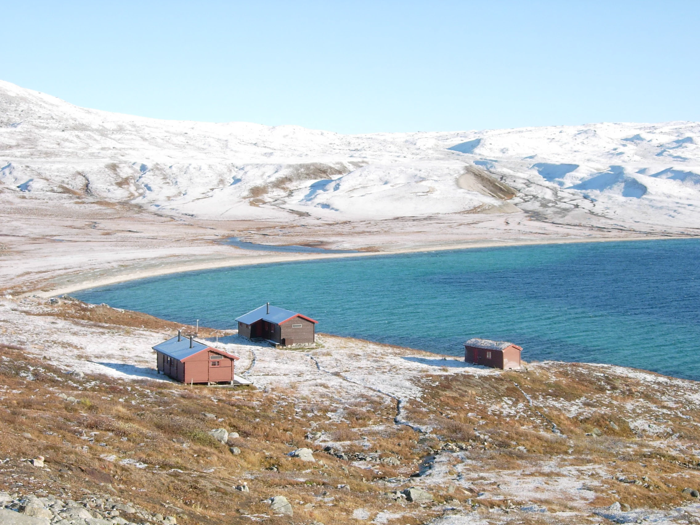 Three small mountain huts on a rocky hillside overlooking a turquoise lake with snow-covered mountains