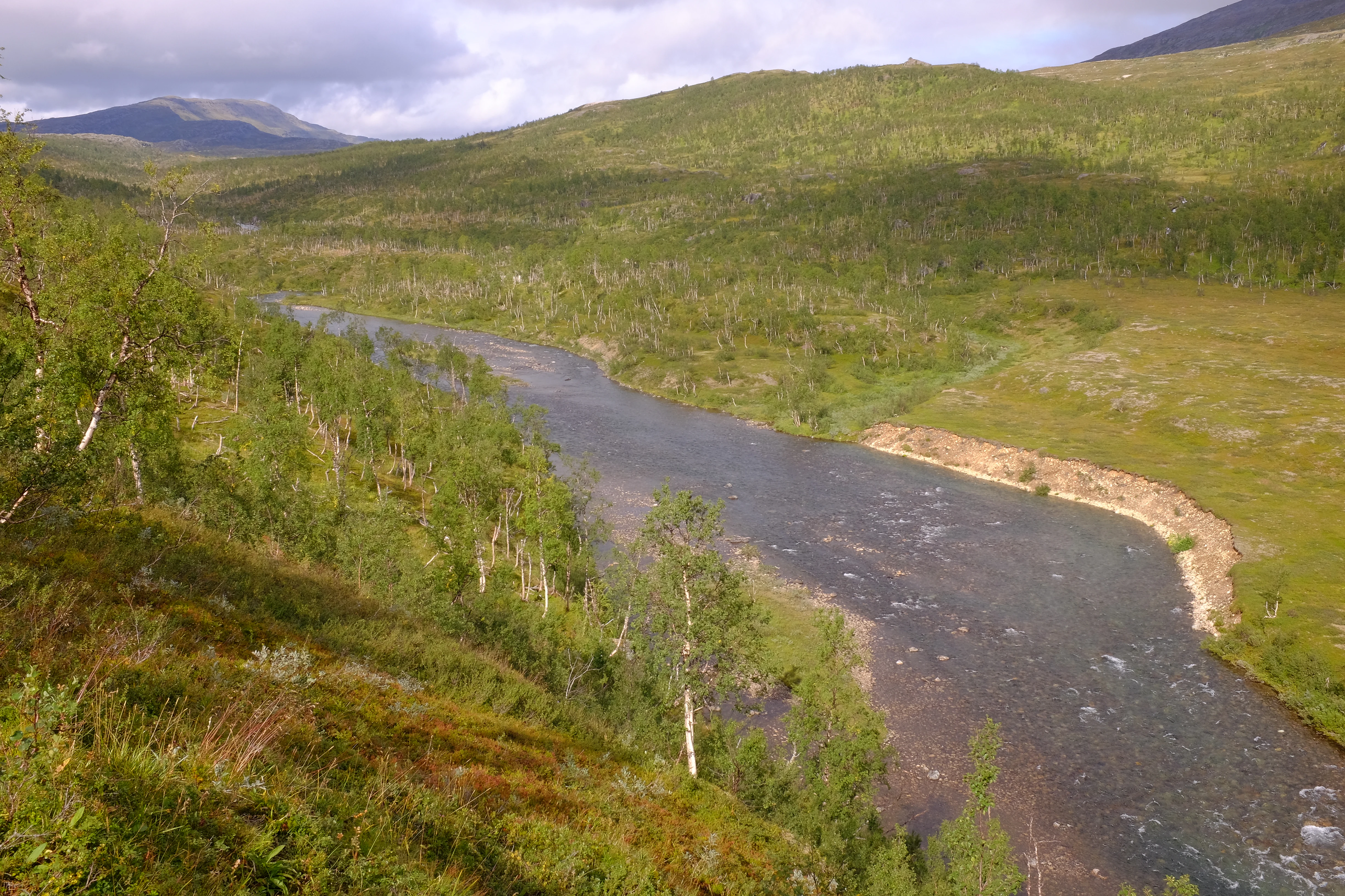 A river flows through a valley surrounded by green hills and trees under a partly cloudy sky