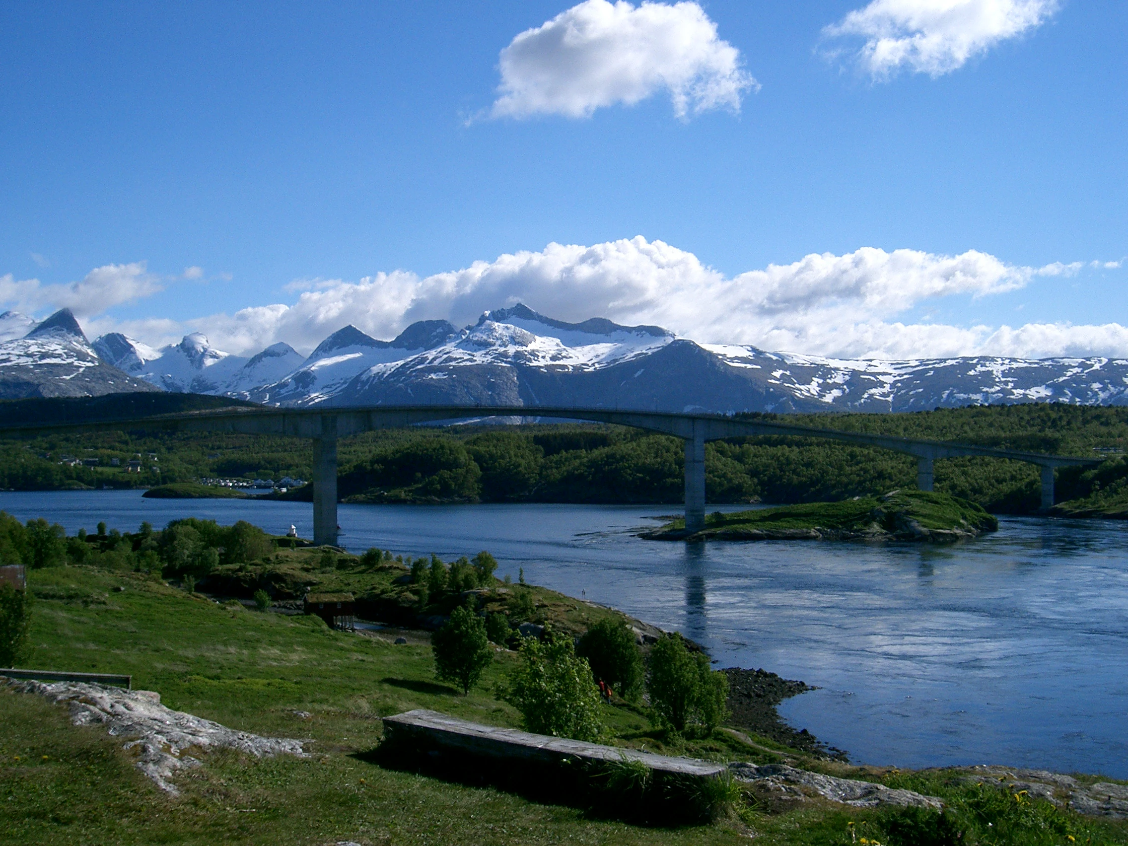 Una toma amplia del estrecho de marea de Saltstraumen bajo un cielo azul, con un largo puente de hormigón que se extiende sobre el agua, picos montañosos nevados en la distancia y hierba verde en primer plano.