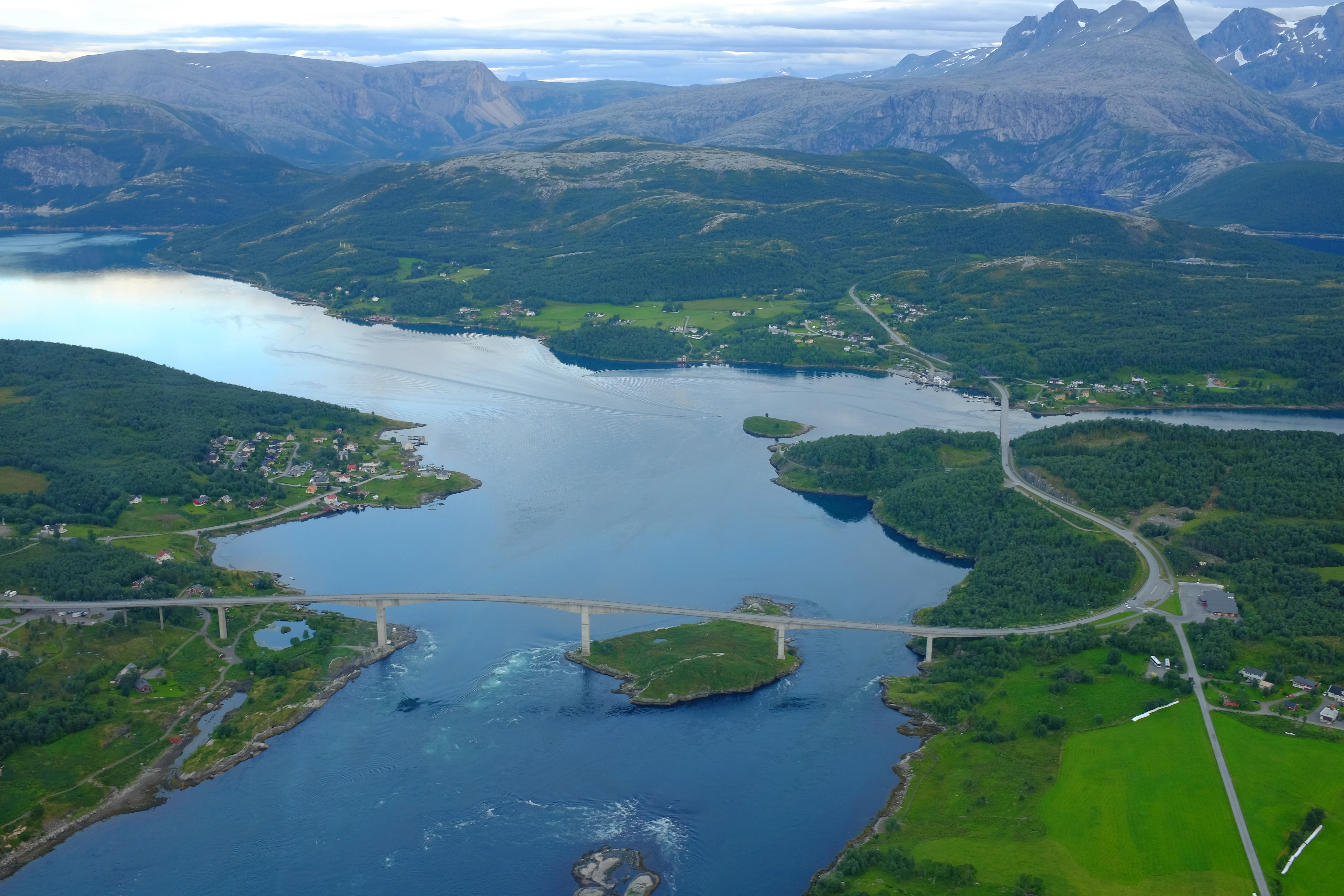 Una vista aérea del estrecho de marea Saltstraumen en Noruega, con un largo puente que cruza el agua, rodeado de colinas verdes, bosques y montañas lejanas.