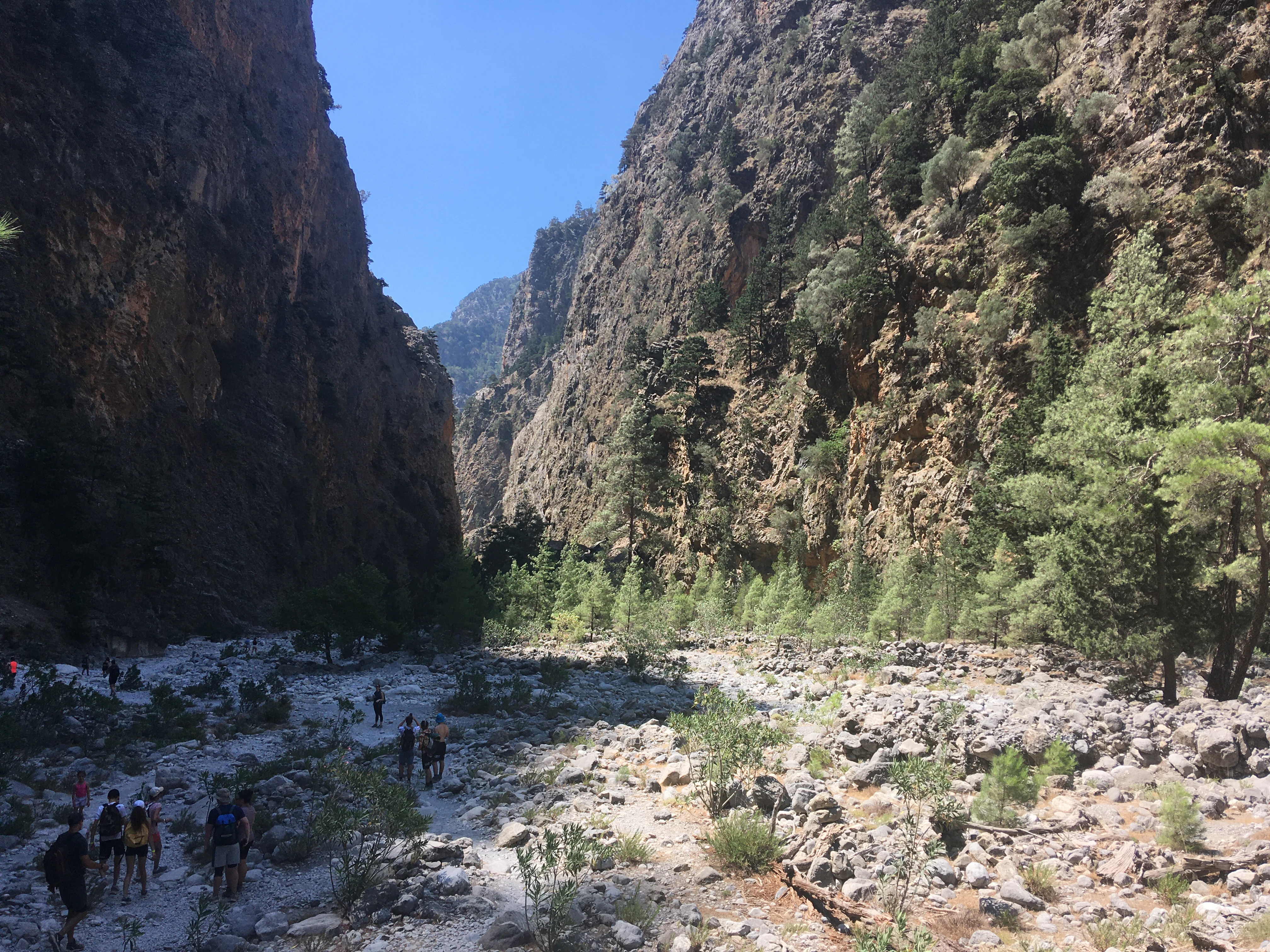 Hikers walk along a wide, stony riverbed at the bottom of a deep, narrow canyon with steep, towering rock walls and sparse trees.