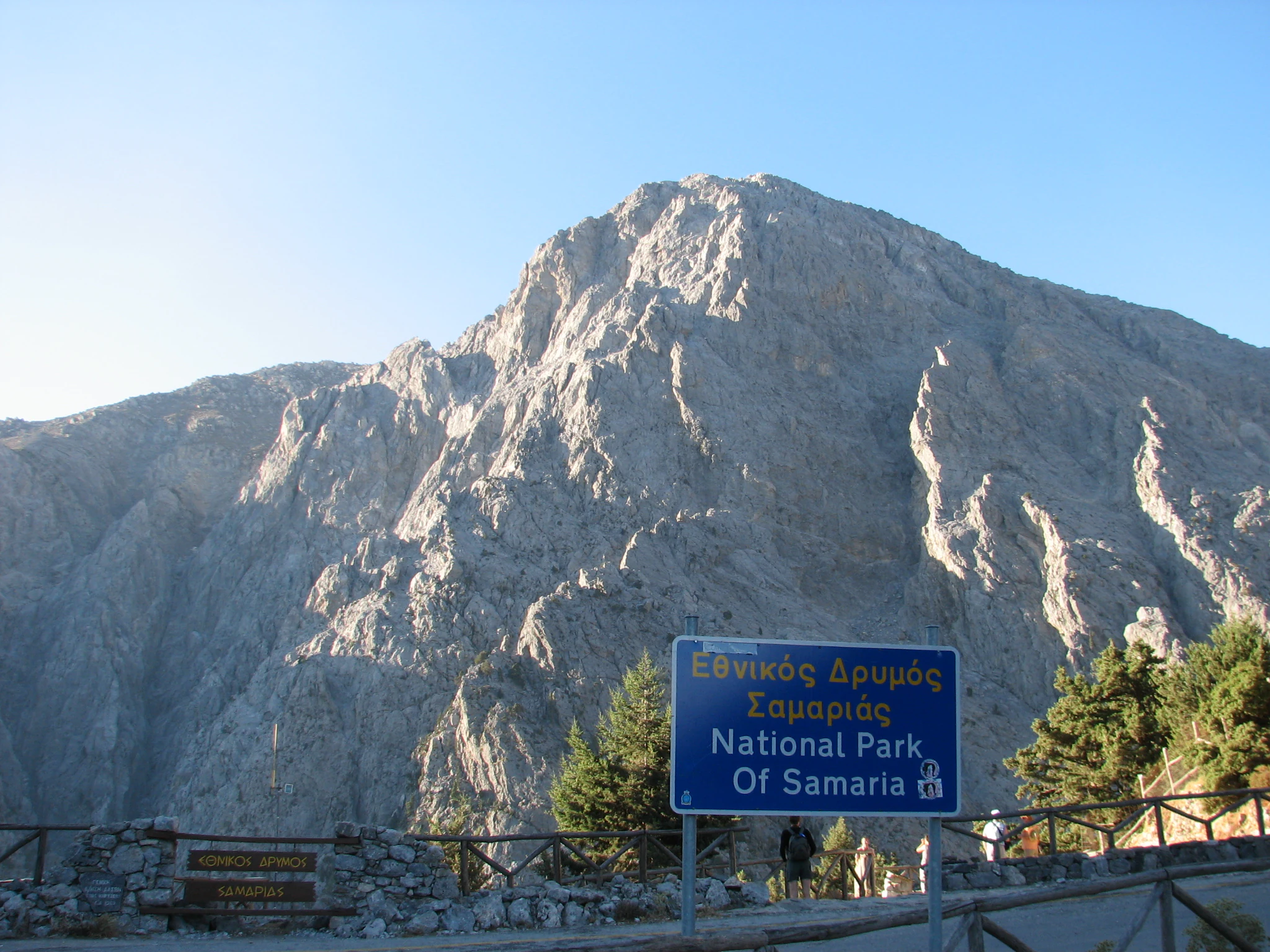 A blue sign reading National Park of Samaria in Greek and English sits in front of a large, rocky mountain peak.