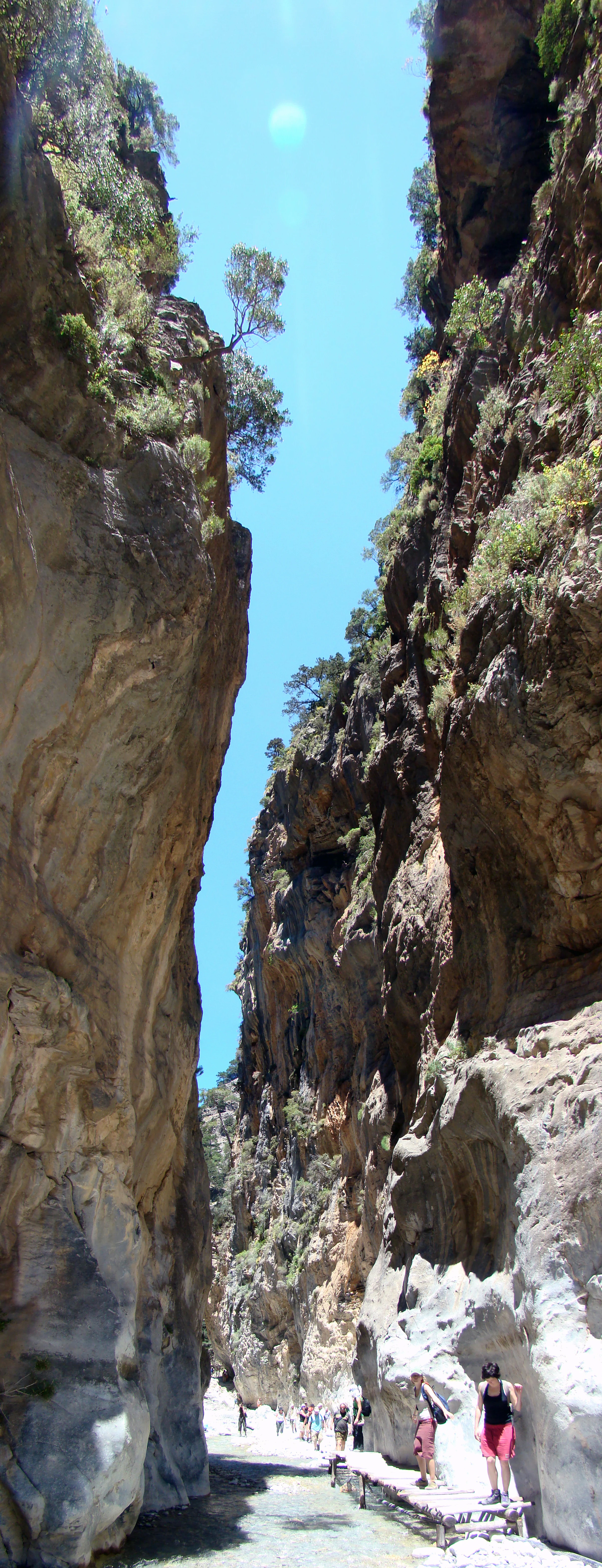 A low-angle view looking up at the high, towering vertical rock cliffs of Samaria Gorge in Samaria National Park, with a group of hikers walking along a wooden path at the base of the gorge.