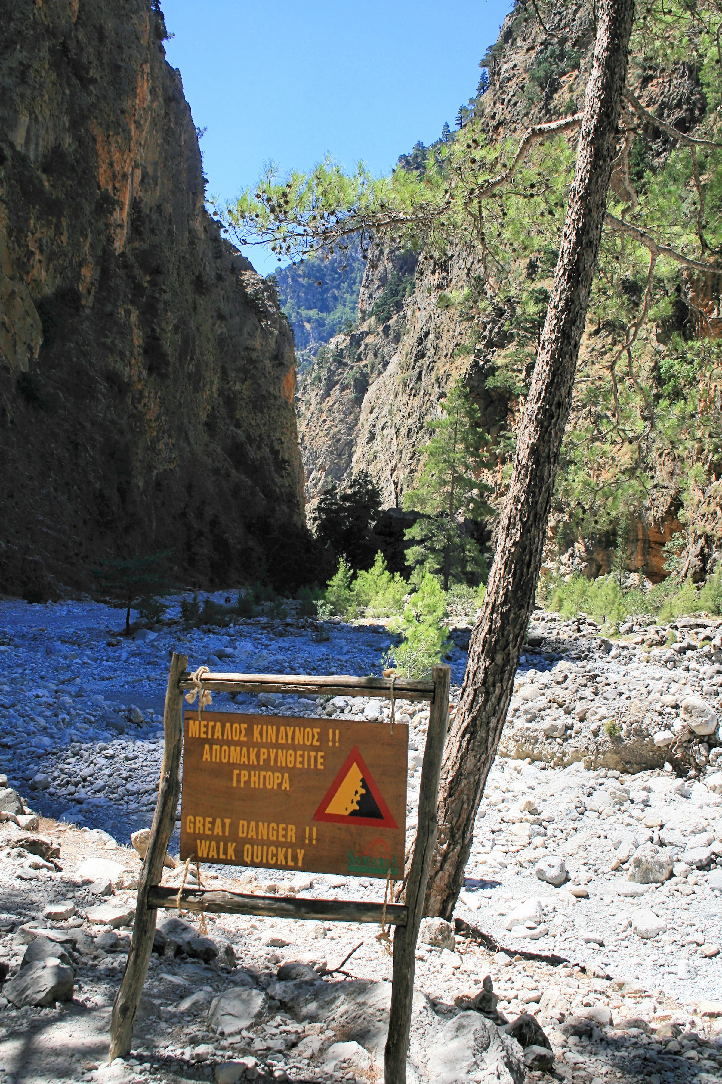 A rustic wooden sign in a rocky canyon warns hikers in Greek and English to walk quickly due to the danger of rockfall.