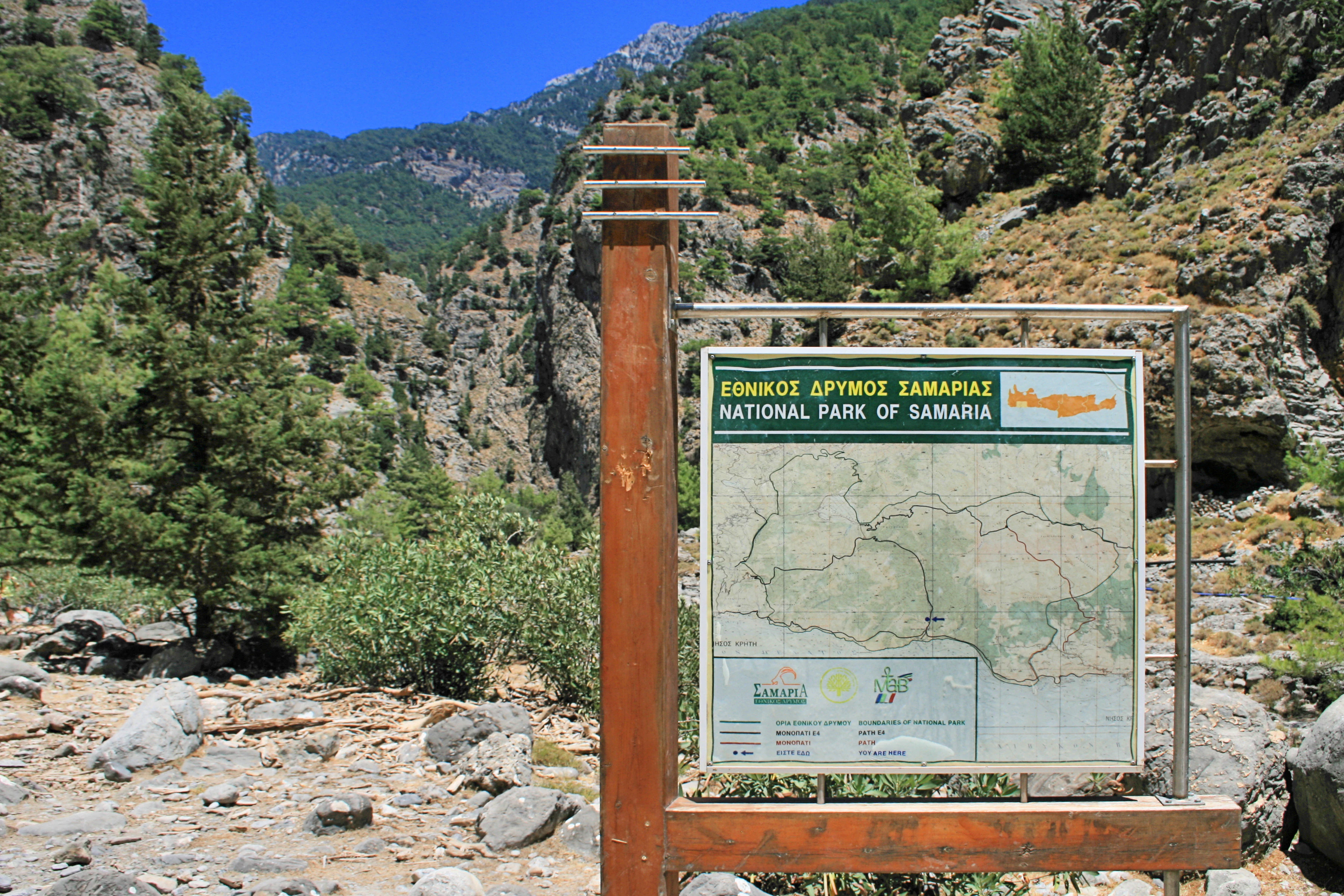 A square metal and wood park sign displaying a map of the Samaria National Park area, set against a backdrop of steep rocky gorge walls and greenery.