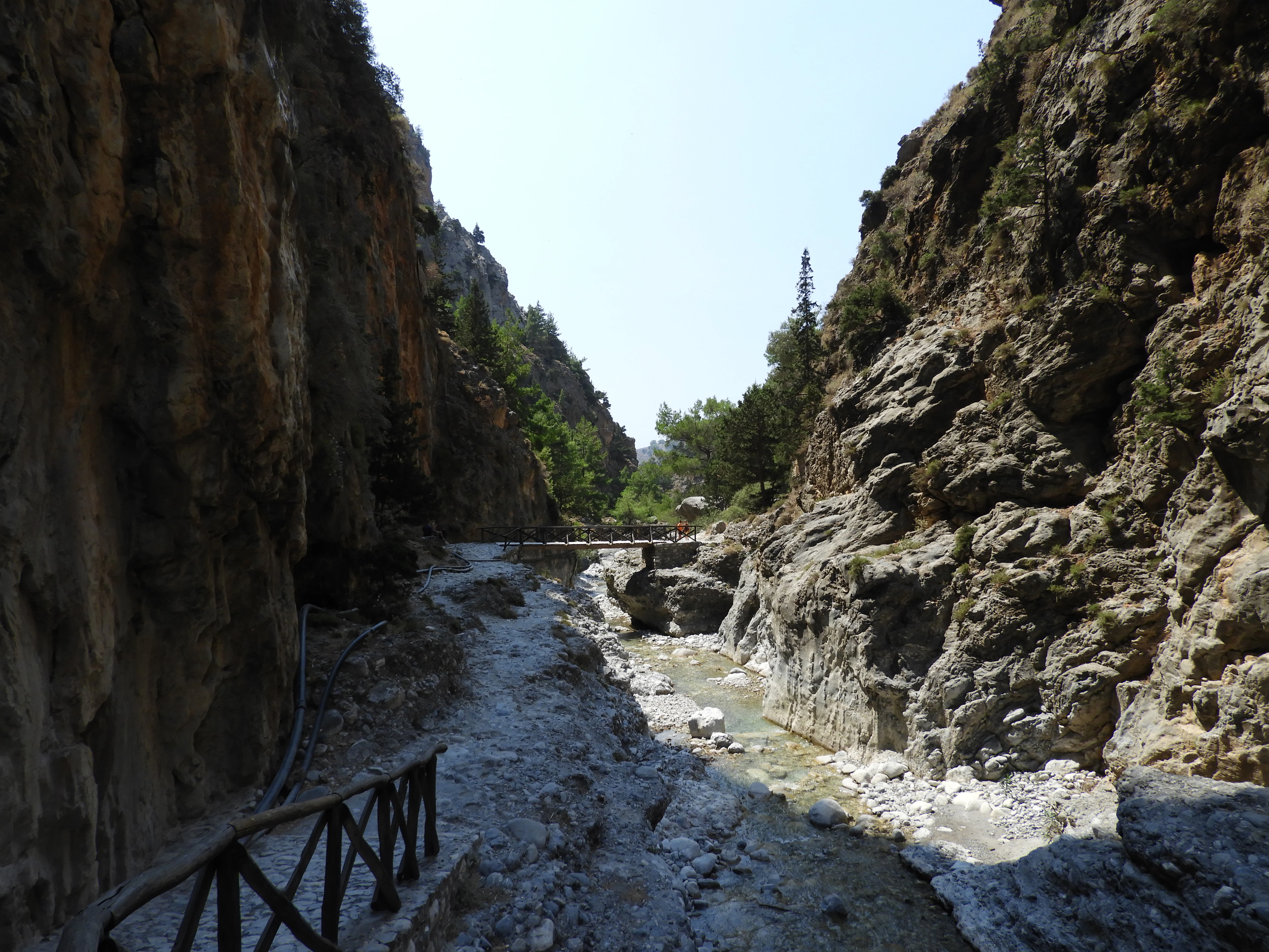 A narrow gravel trail runs alongside a small stream within a deep, steep-walled limestone gorge, with a wooden railing in the foreground.