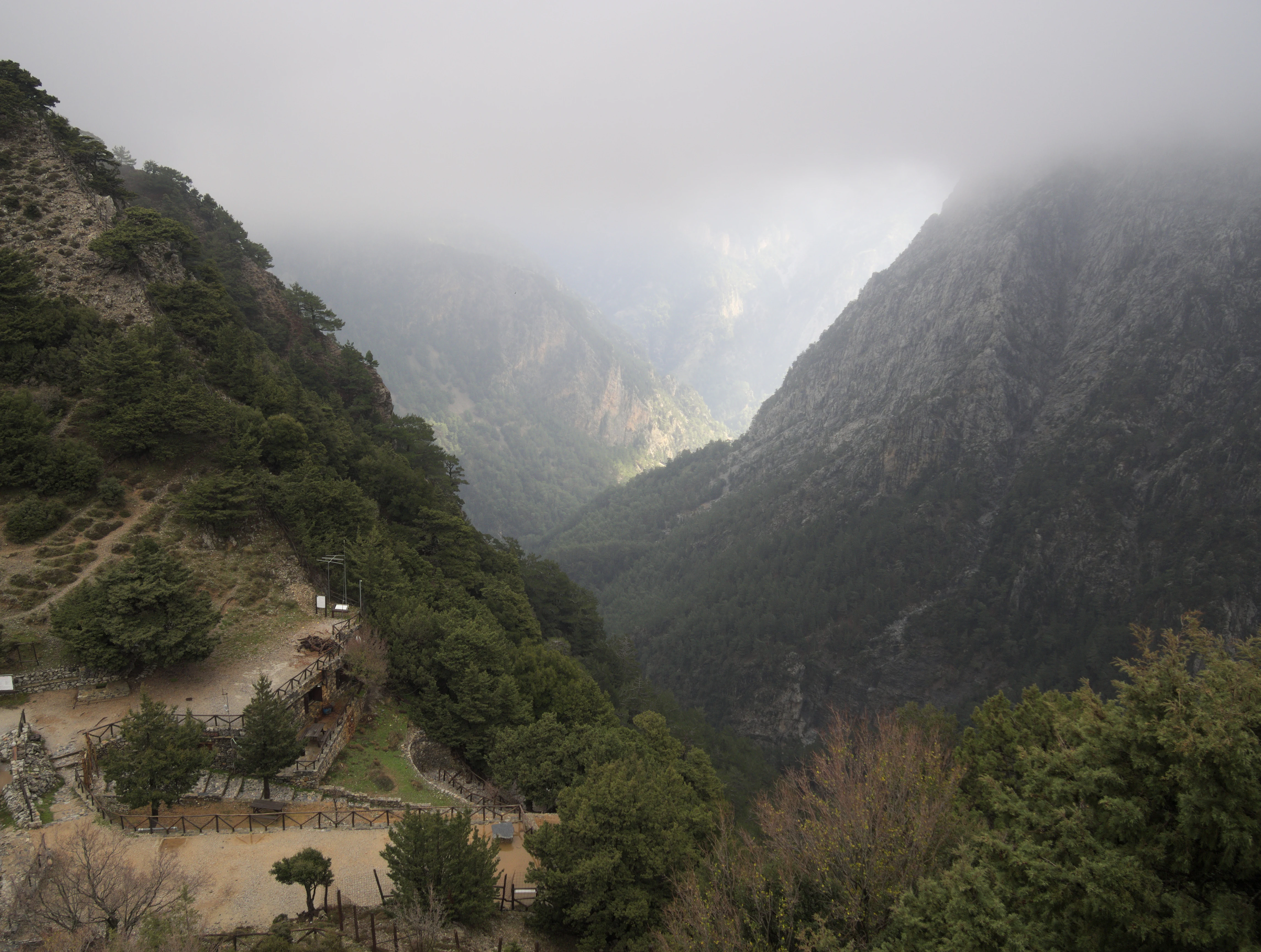 A wide, elevated view of a deep mountain gorge on a foggy, overcast day. The foreground shows a dirt path and sparse vegetation on a slope, leading the eye into a steep, mist-filled canyon.