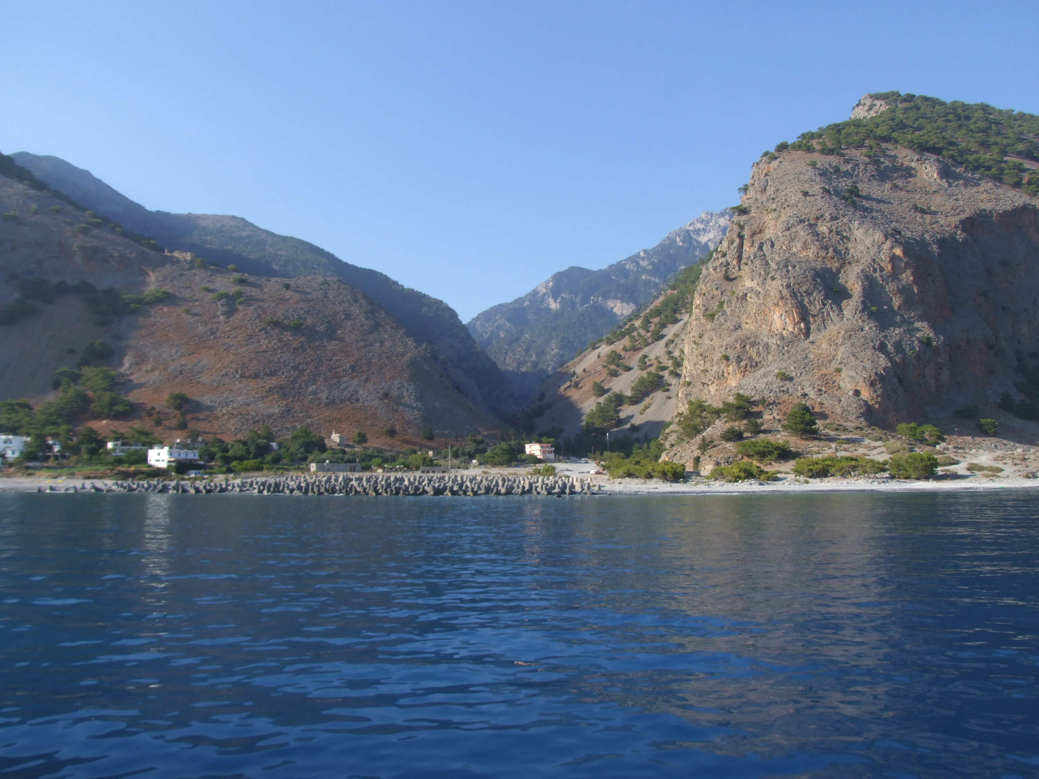 A wide angle view from the sea looking toward the coast of Agia Roumeli, where the Samaria Gorge meets the Libyan Sea, featuring steep rocky cliffs and a pebble beach.