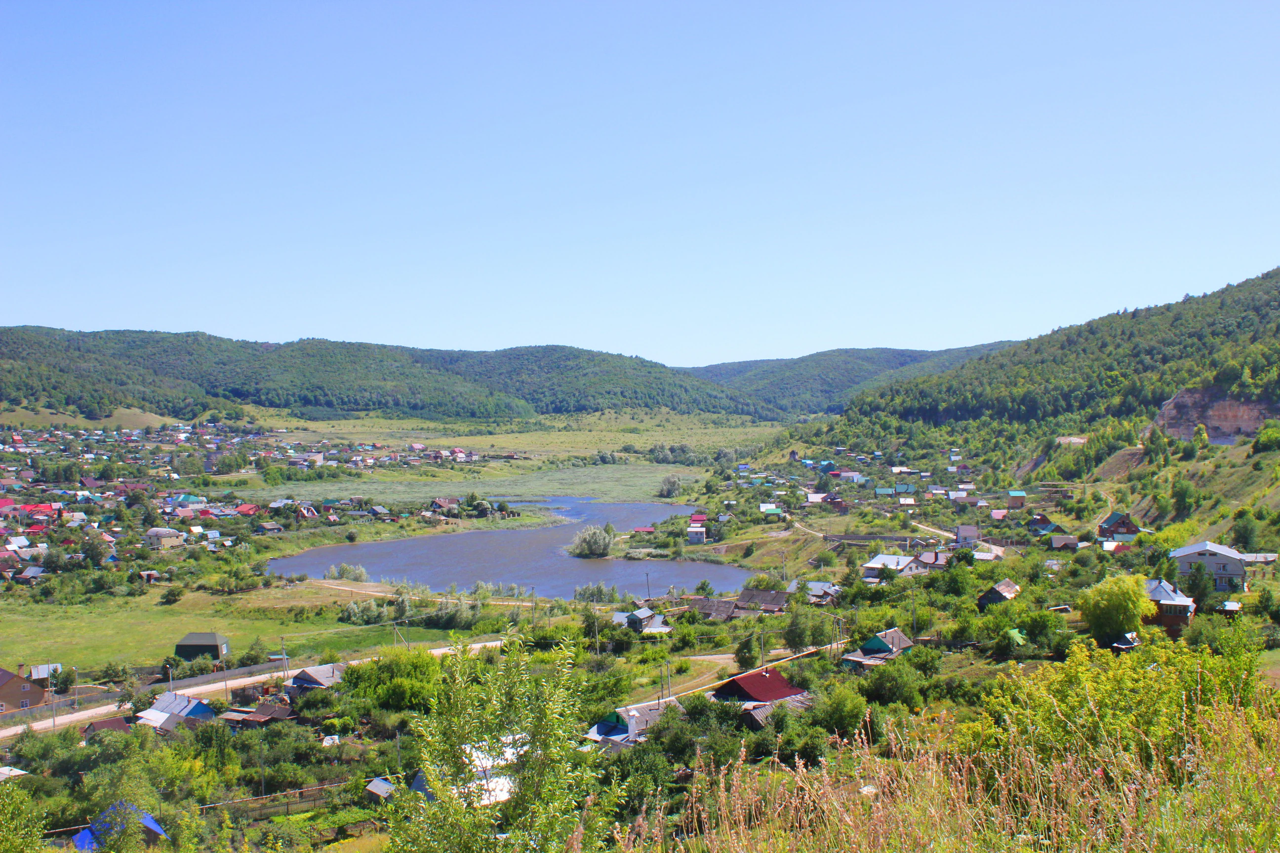 A high-angle panoramic view of a river valley containing a small village, lush green hills, and a winding body of water under a clear blue sky.