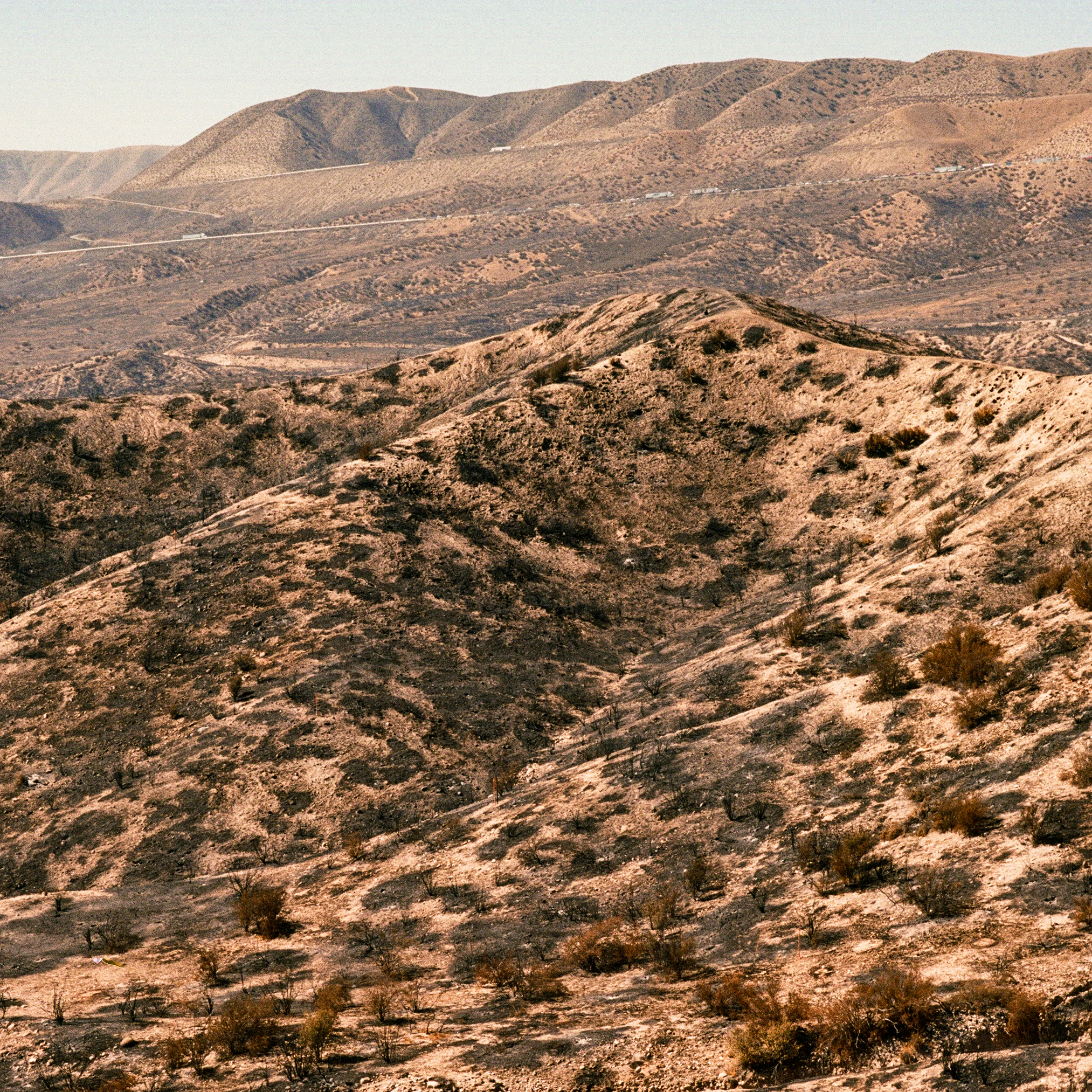 A wide, arid landscape of rolling hills and slopes showing evidence of fire damage, with blackened vegetation and exposed dry earth under a clear sky.