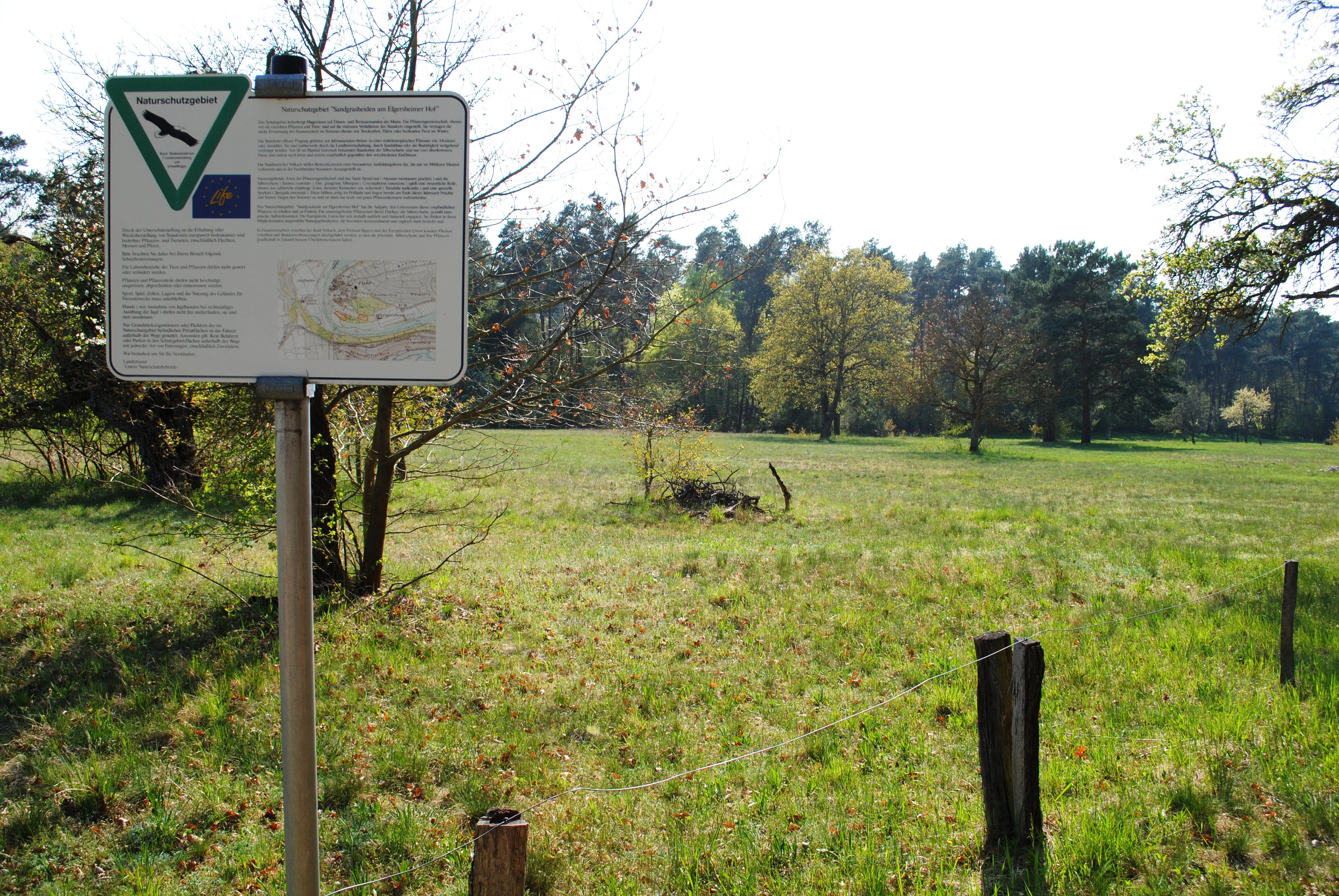 A metal information sign on a post in the foreground, with an open grassy field and forest in the background.