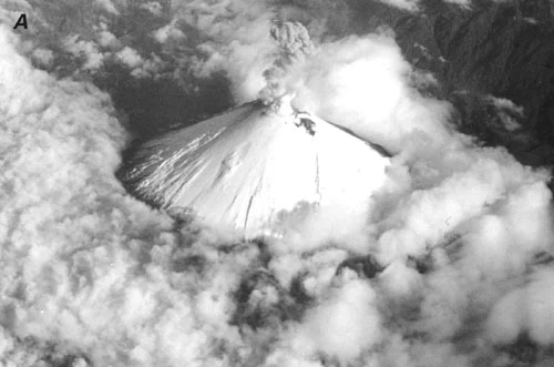 Aerial view of a snow-capped volcano with an ash plume rising from the summit, surrounded by clouds
