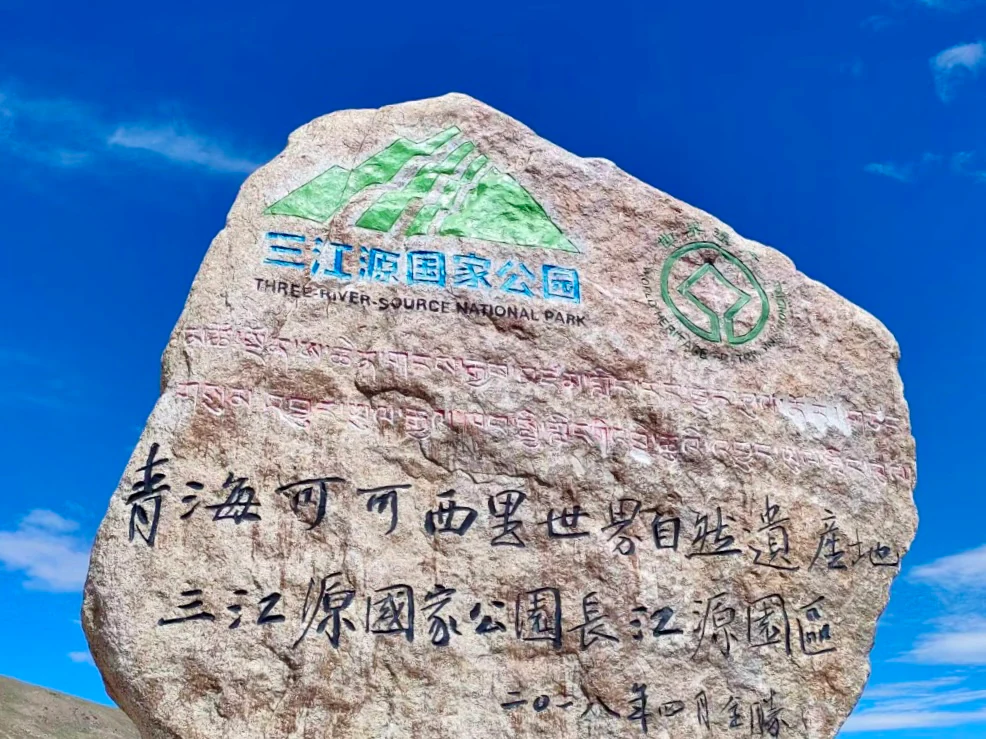 Marcador de piedra con texto en chino y traducción al inglés para el Parque Nacional de la Fuente de los Tres Ríos y el sitio del Patrimonio Mundial de Qinghai Kekexili, con cielo azul.