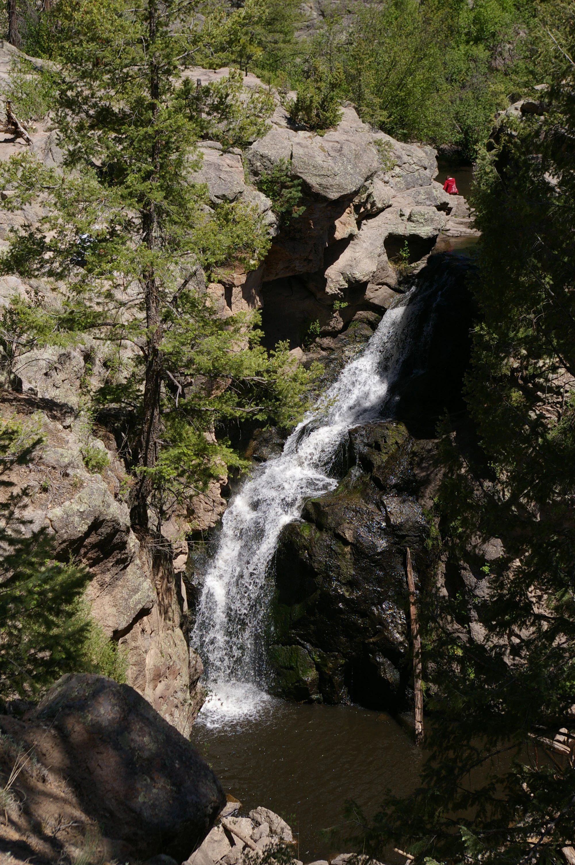 A vertical view of Jemez Falls dropping over dark rocky cliffs into a pool, framed by pine trees and rugged terrain.