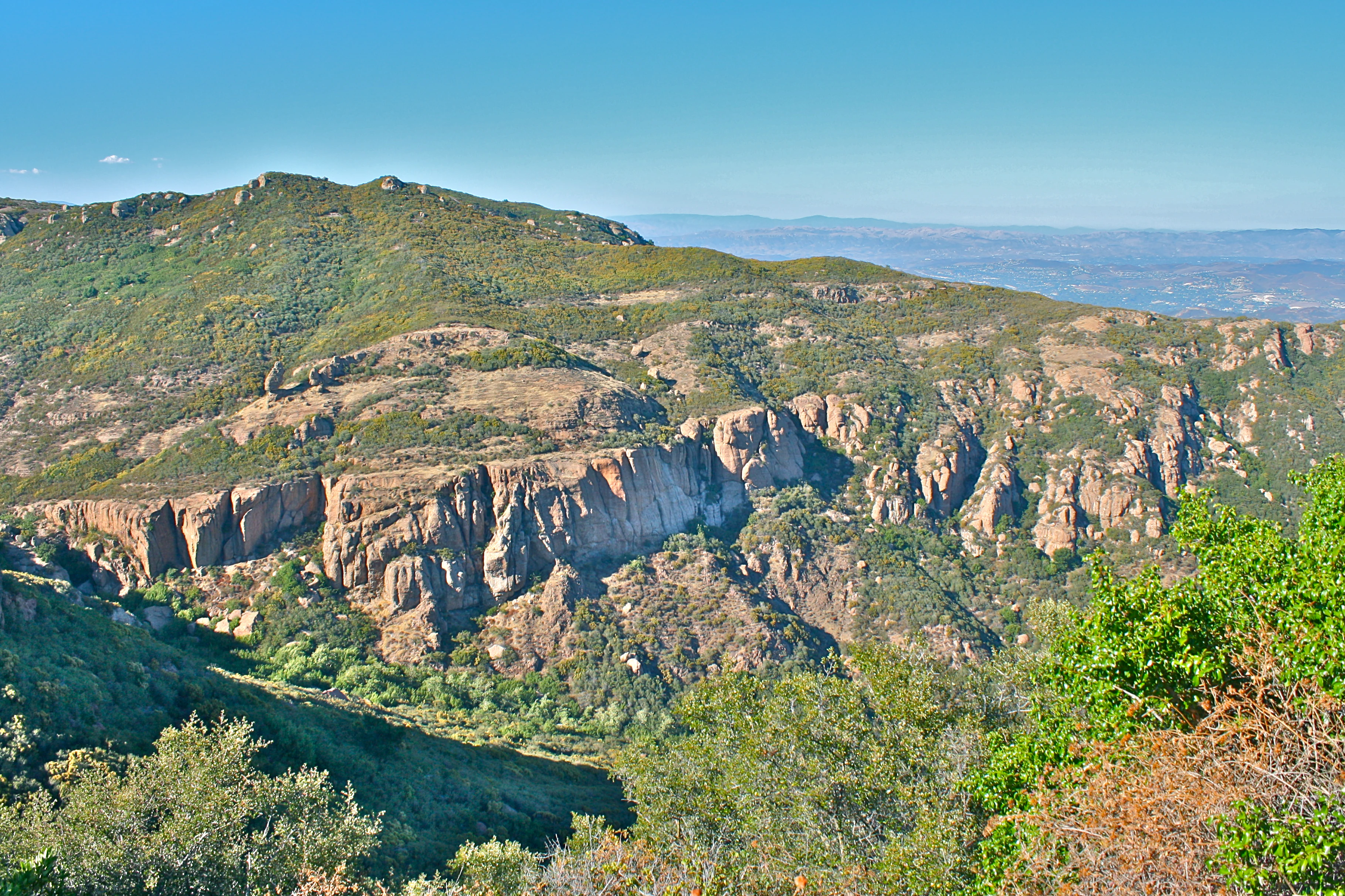 A wide landscape view of rocky sandstone cliffs and green hills under a clear blue sky, with a hazy valley visible in the background.