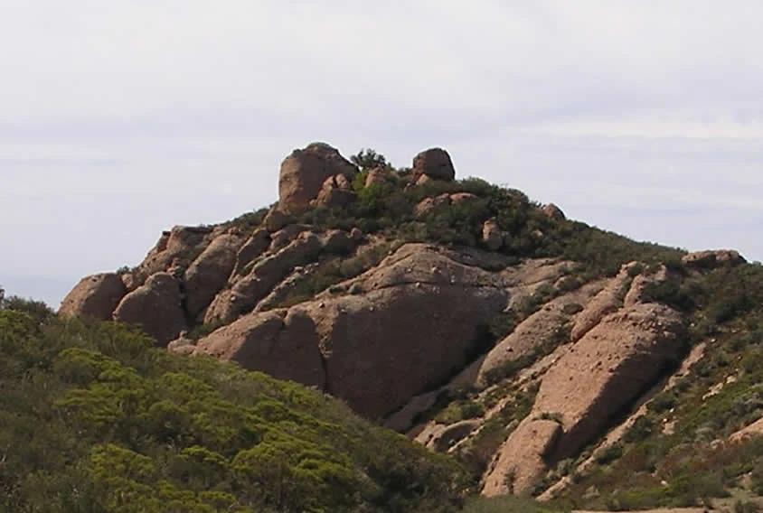 A rocky mountain peak with large boulders and green shrubs under a cloudy sky.