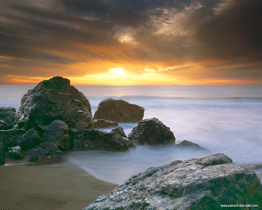 Large dark rocks sit on a sandy beach with smooth misty water and a dramatic orange and grey sunset sky.
