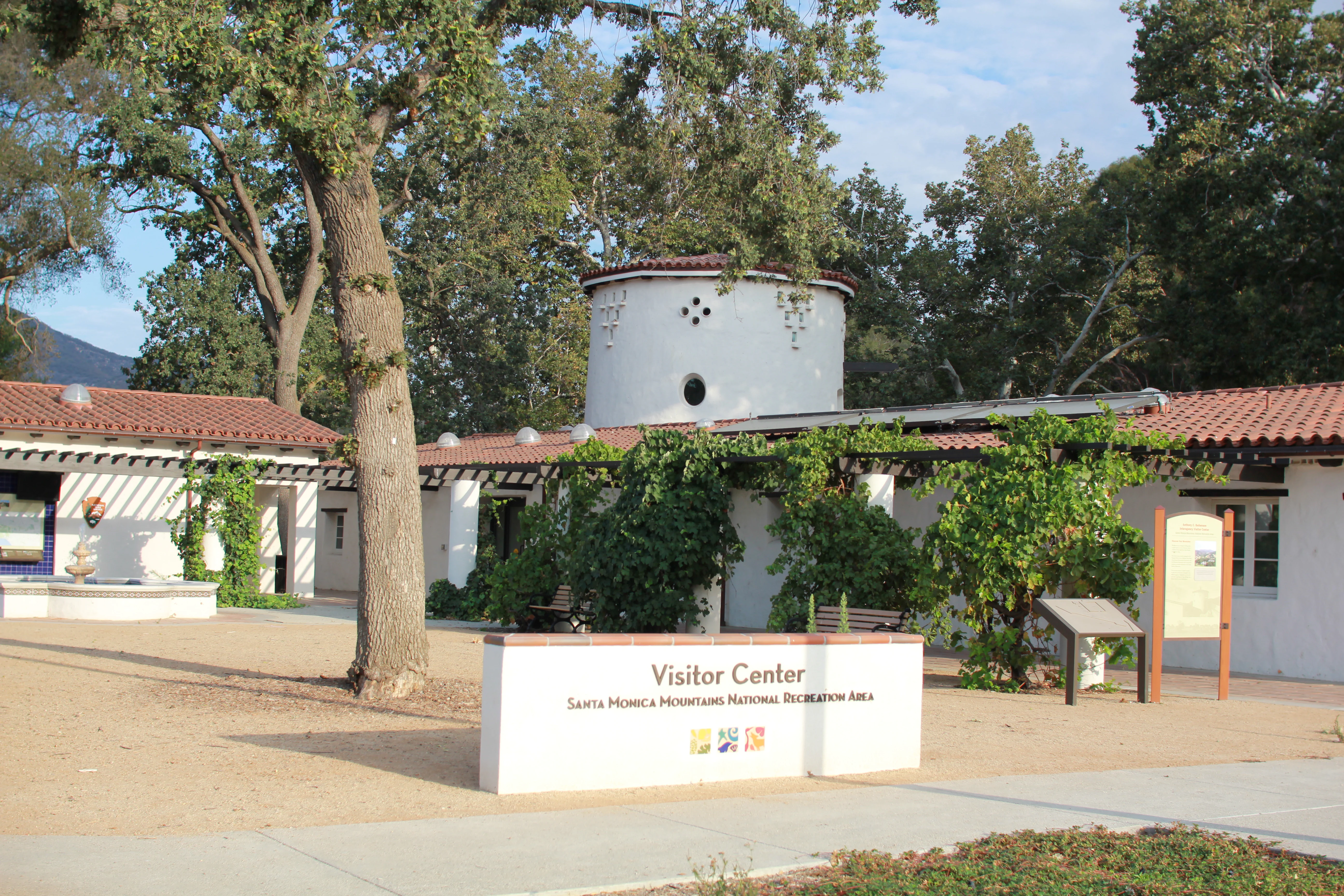 A white single-story building with a red tile roof and a cylindrical tower, featuring a large white sign in the foreground reading 'Visitor Center Santa Monica Mountains National Recreation Area'.