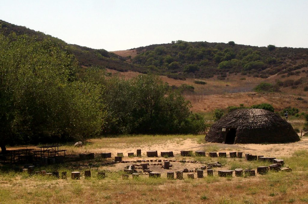 A landscape view showing a traditional Native American dome-shaped structure made of natural materials alongside a circular arrangement of wooden post stumps, with chaparral-covered hills in the background