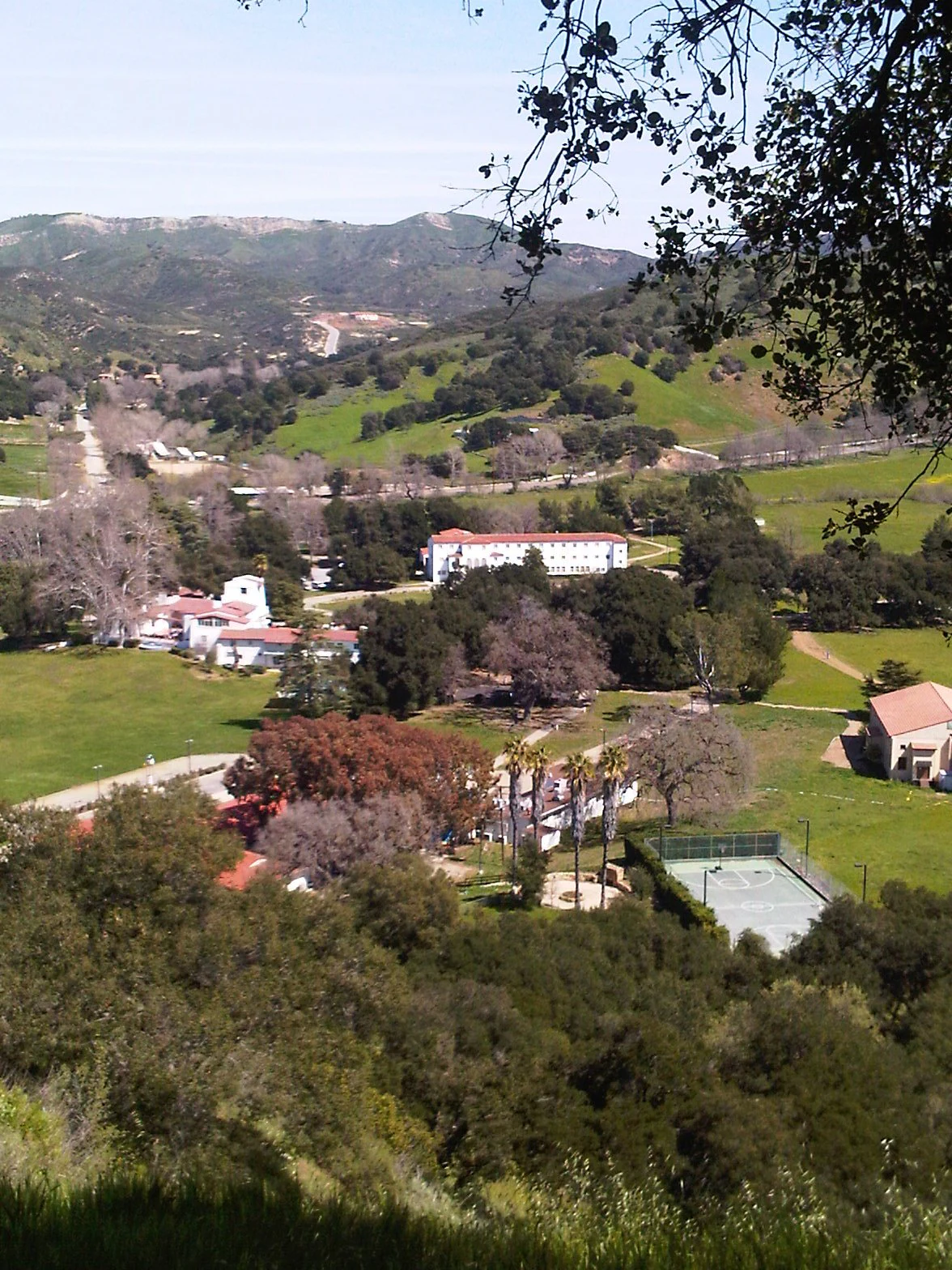 A wide landscape view showing white buildings with red roofs, a tennis court, and palm trees situated in a green valley surrounded by rolling hills and mountains.