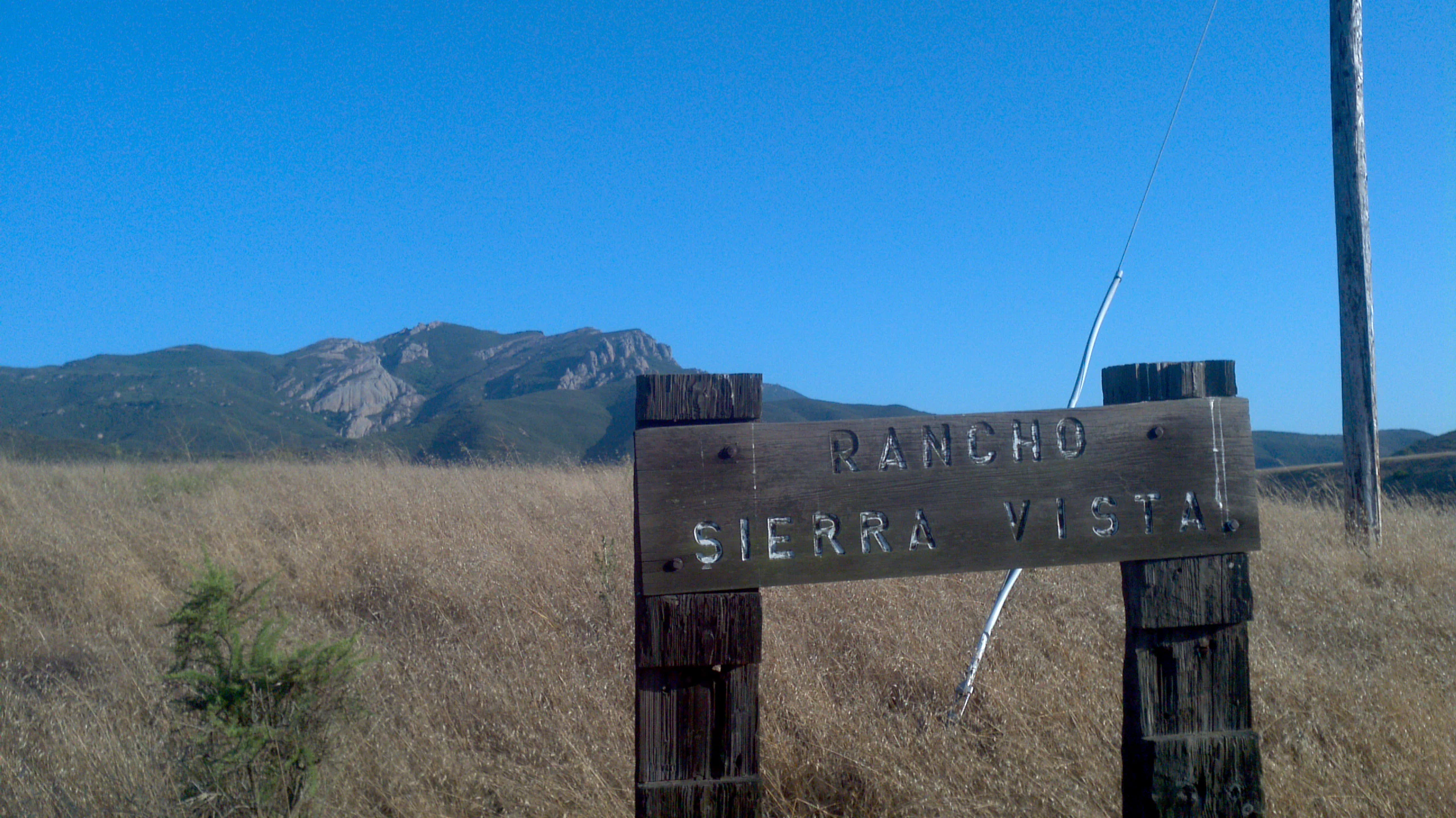 A weathered wooden sign reading 'RANCHO SIERRA VISTA' mounted on two posts in a dry grassy field, with a mountain ridge and clear blue sky in the background.