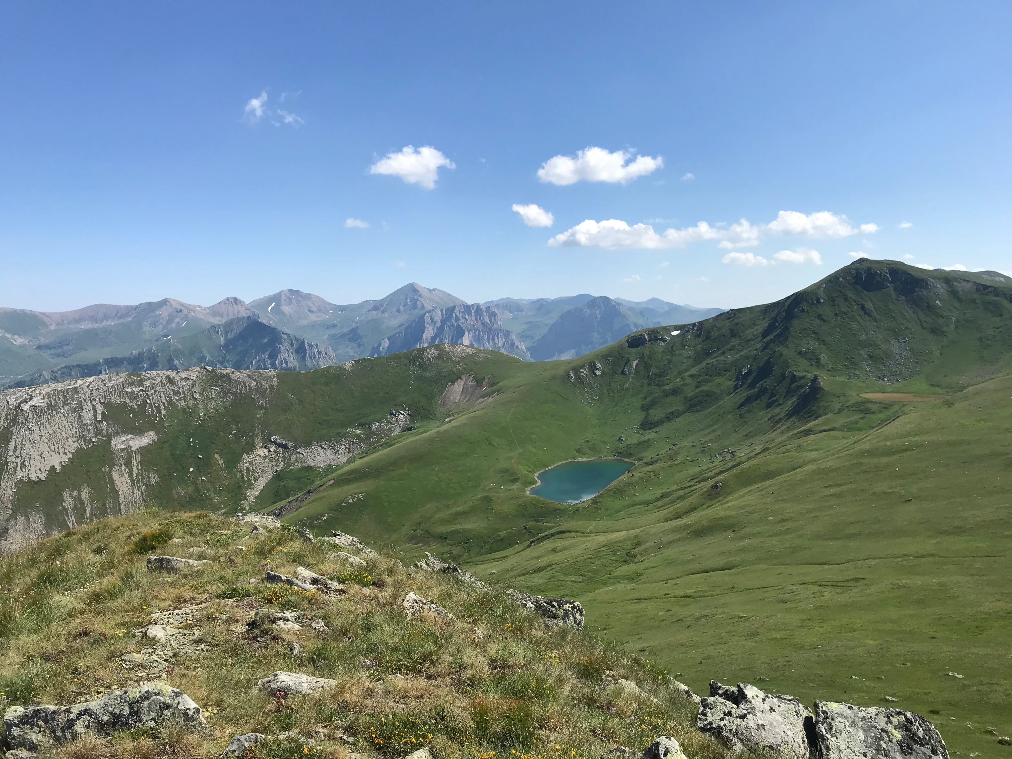 Grassy mountain slope with rocky outcrops, a small lake in the valley, and distant mountain peaks under a clear blue sky with scattered clouds