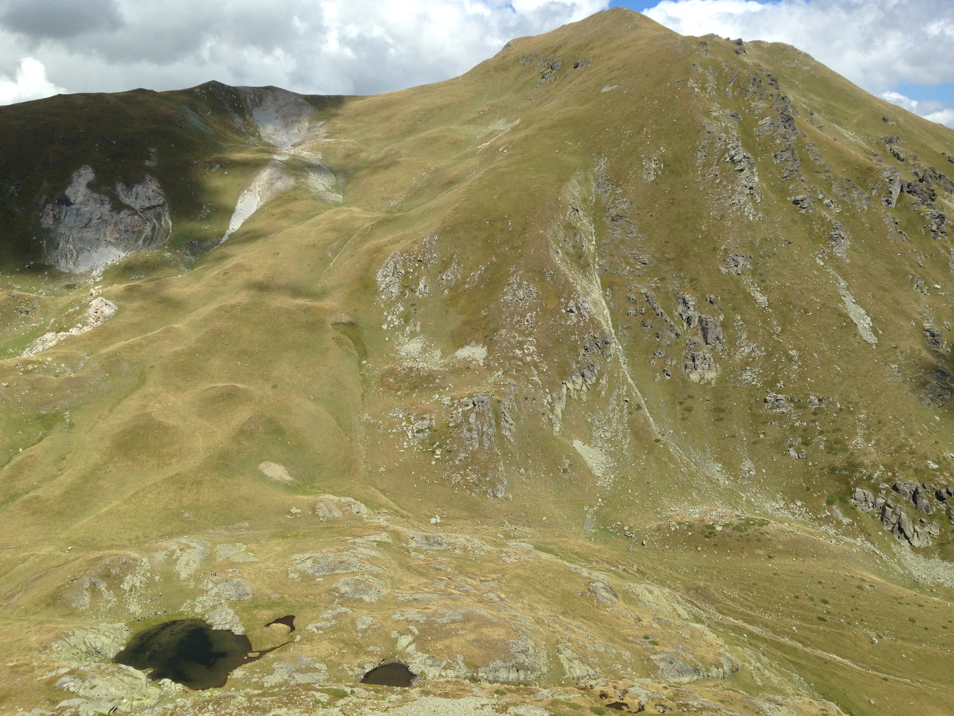 Grassy mountain slope with a small lake in the foreground under a partly cloudy sky